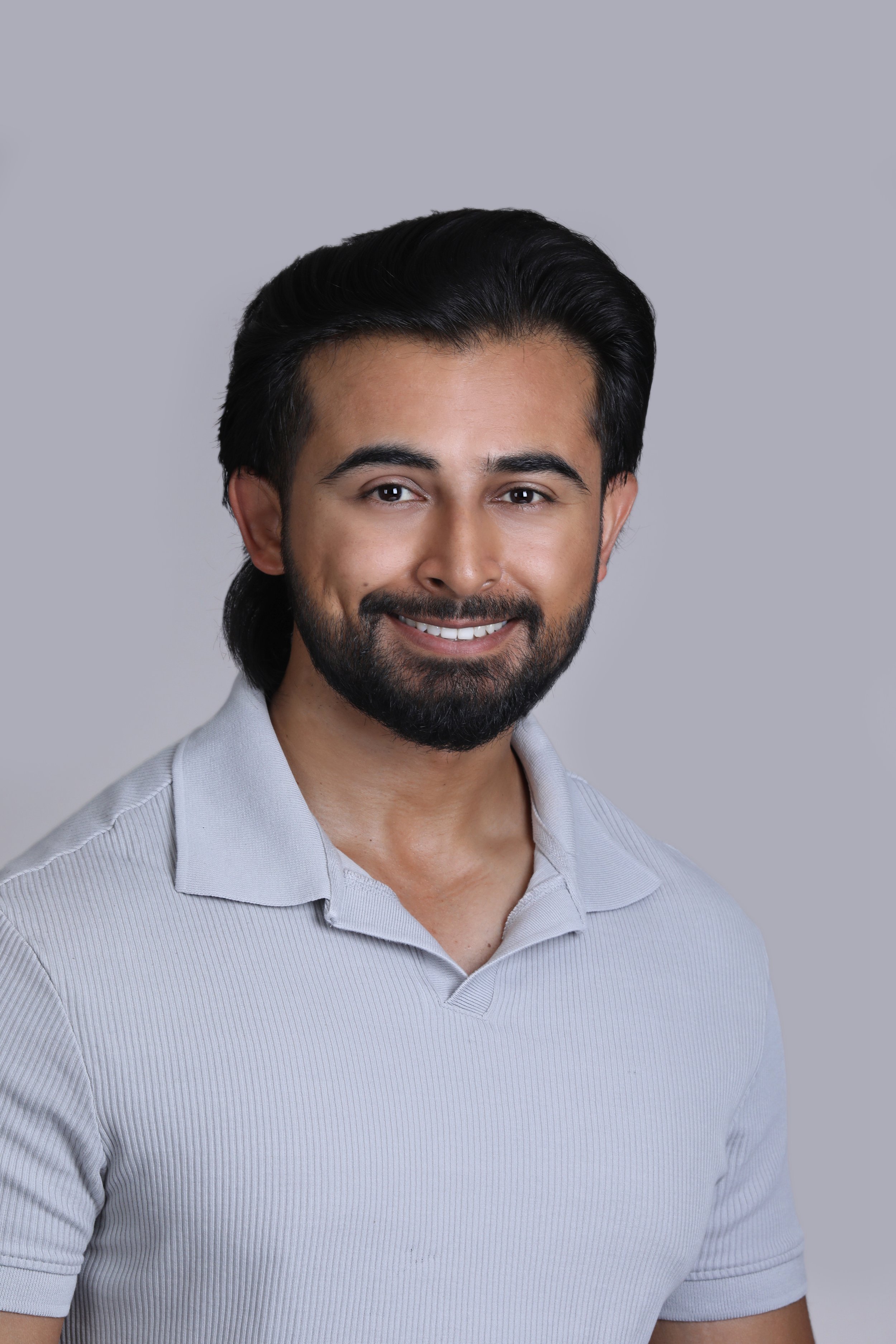 A smiling man with dark hair and a beard wearing a light gray polo shirt, standing against a plain gray background.