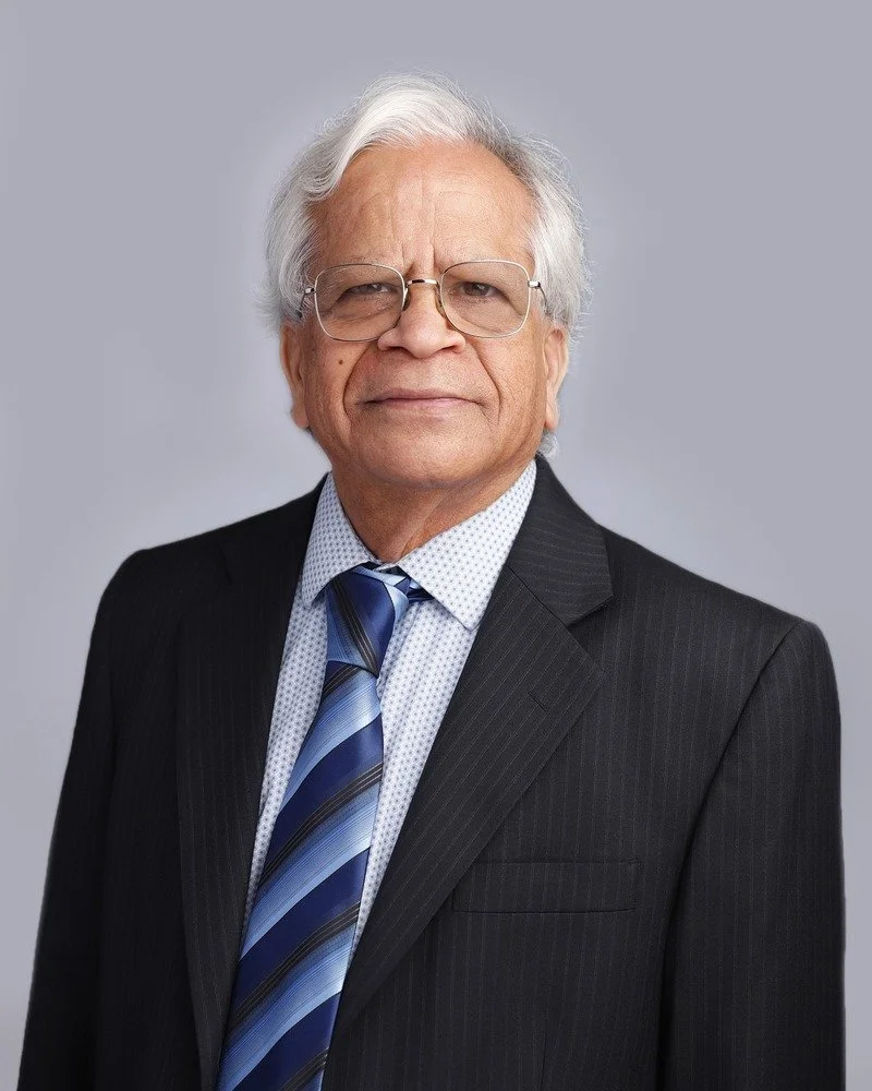 An older man with white hair, glasses, and a serious expression, dressed in a dark pinstripe suit and a blue striped tie, against a plain gray background.