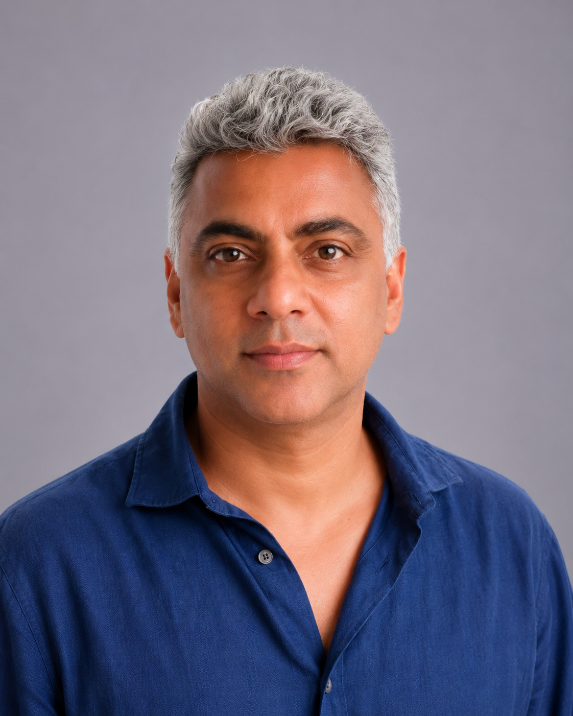 Headshot of a middle-aged man with short, curly gray hair, wearing a navy blue collared shirt, against a neutral gray background.