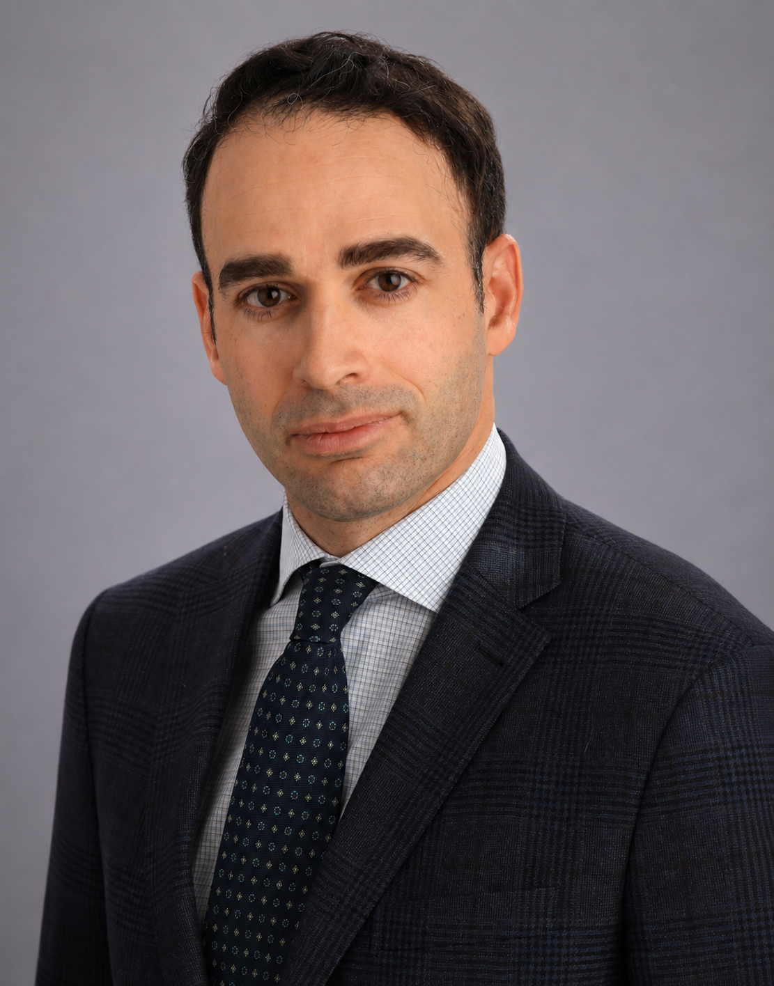 Professional headshot of a man in a dark suit, light shirt, and patterned tie against a plain gray background.
