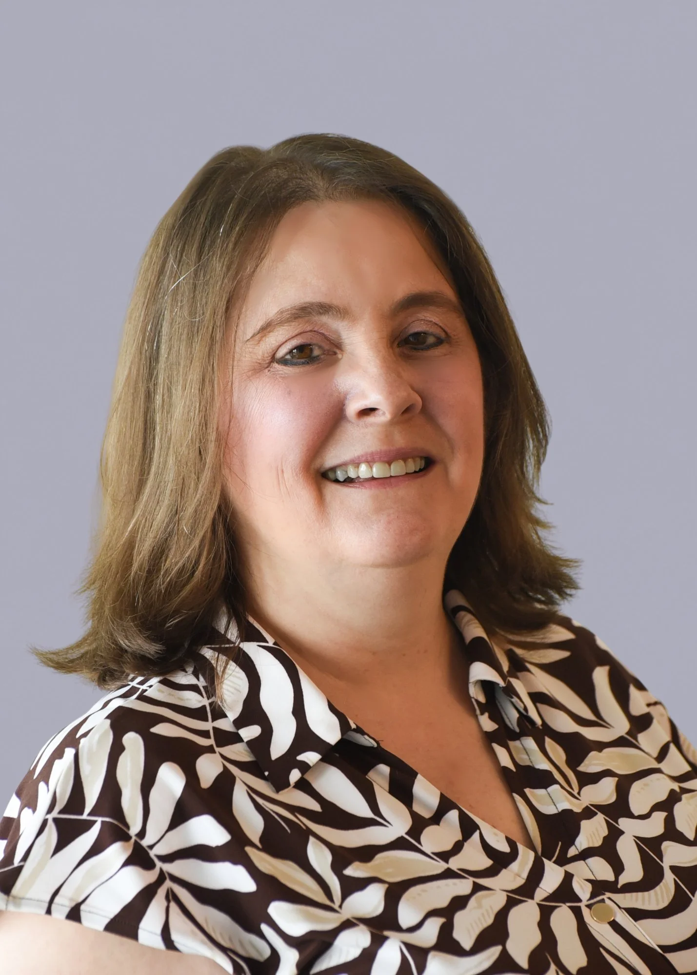 Close-up of a smiling woman with shoulder-length brown hair wearing a black and white patterned button-up shirt, against a light gray background.