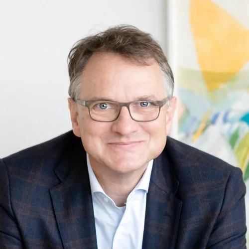 Porträt eines Mannes mit Brille, Hemd und Jacke, lächelnd, im Hintergrund ein farbiges Kunstwerk.