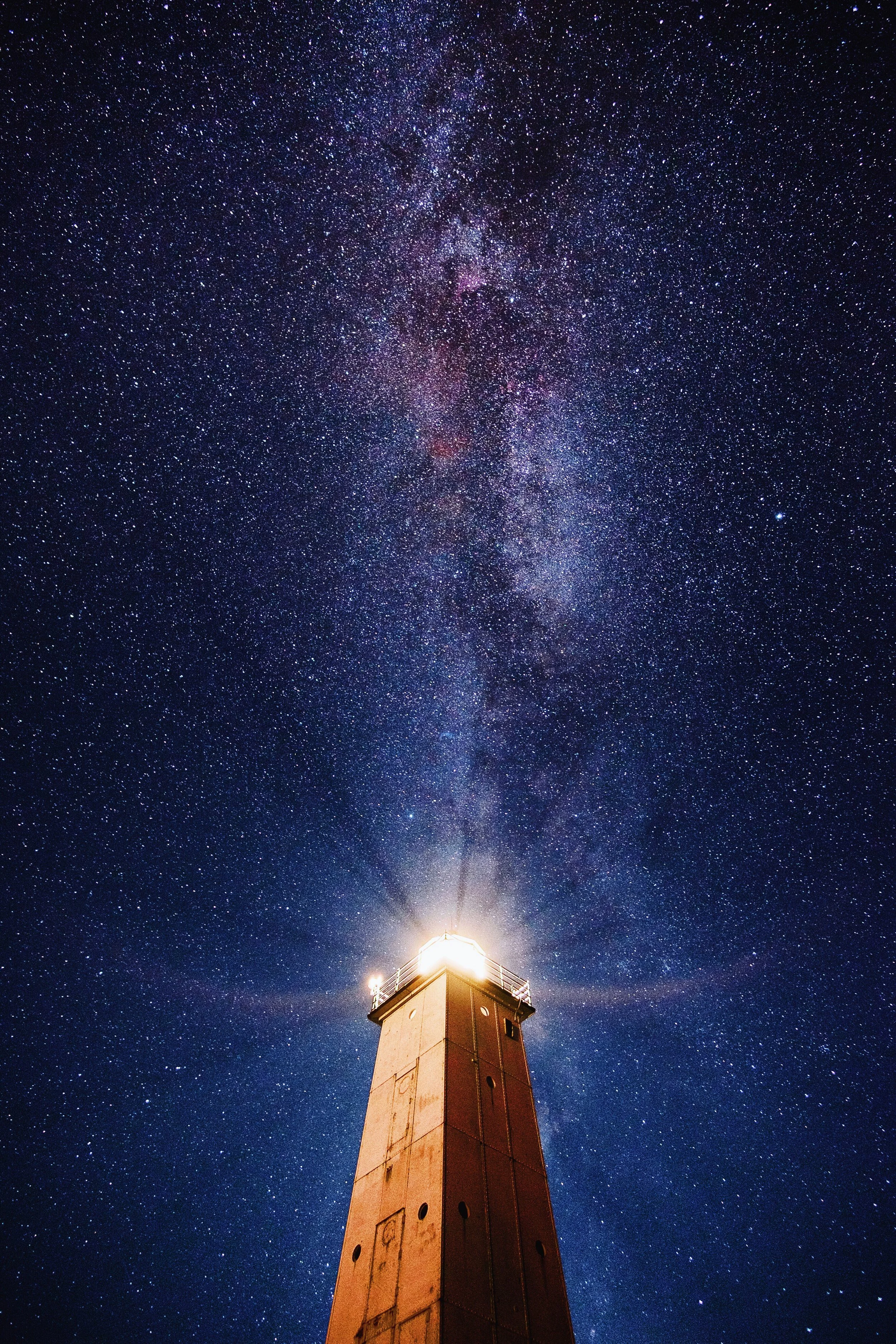 Lighthouse beacon illuminating night sky with visible Milky Way galaxy stretching overhead against deep blue starry background