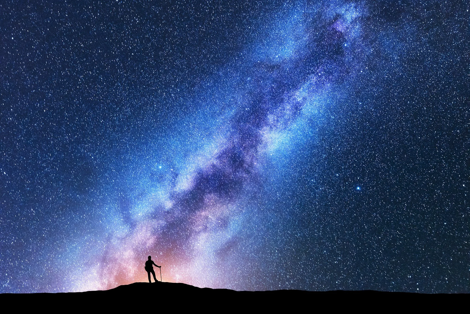 Hiker silhouette standing on hilltop under spectacular blue and purple Milky Way galaxy stretching across night sky