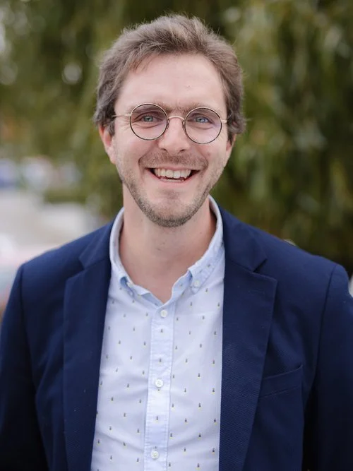 Un homme souriant porte des lunettes rondes, une chemise blanche et une veste bleue, avec un fond de nature