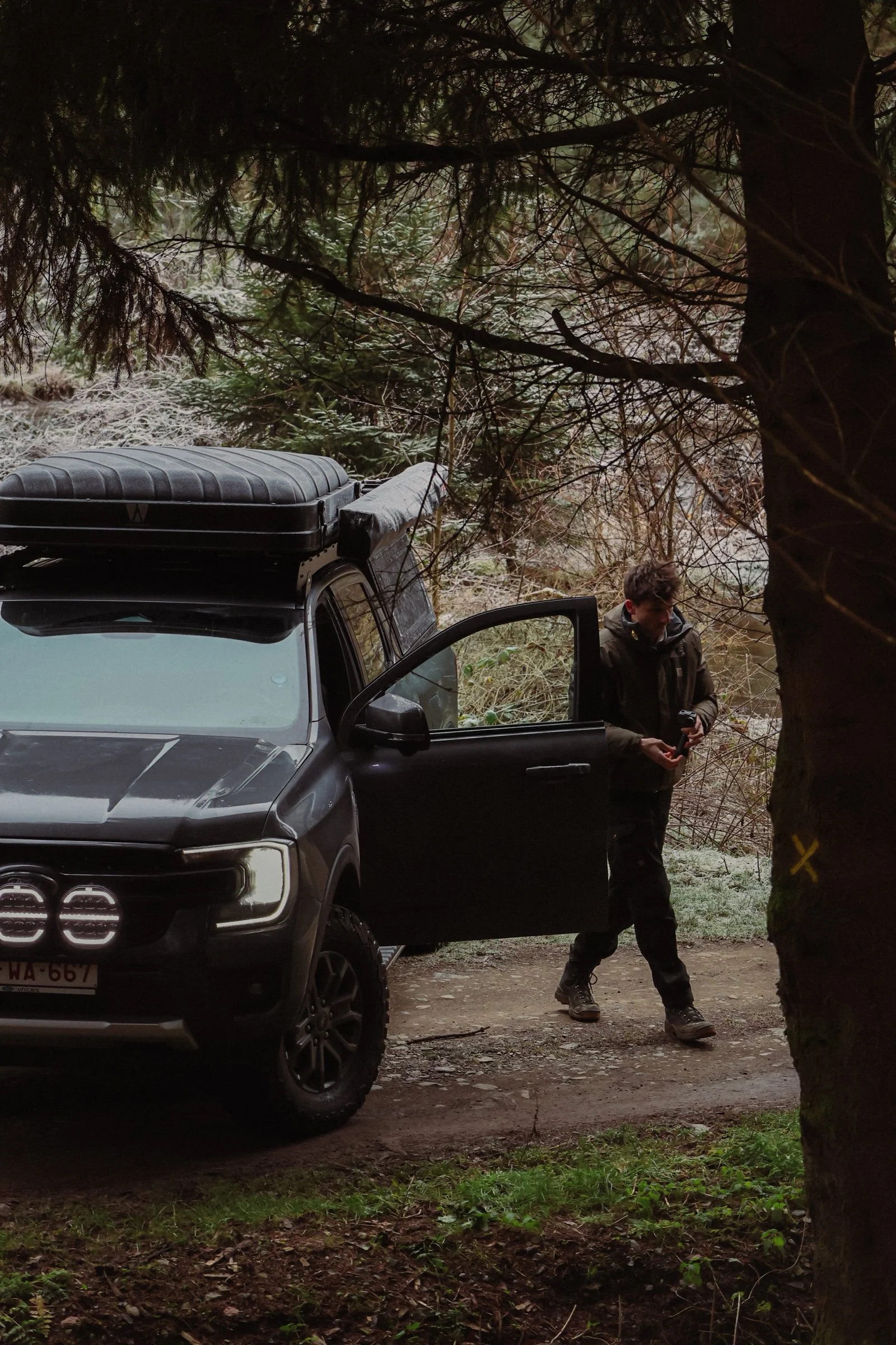 A man with short hair standing outside his black SUV in a forested area, holding a device in his hands, with the car's front door open. The scene appears to be in a wooded, outdoor setting with trees and foliage.