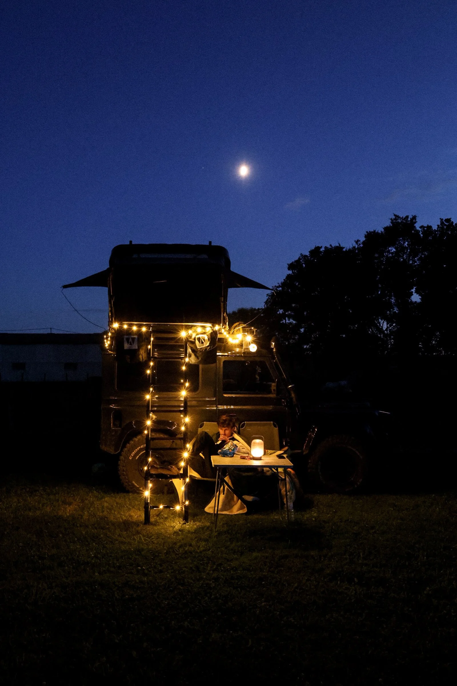 Nighttime camping scene with person sitting by a table, fairy lights on a campervan, and a visible moon in the sky.