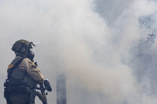 A firefighter wearing protective gear and a helmet in smoky conditions with a helicopter overhead.