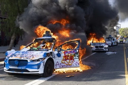 A police car is on fire with thick black smoke billowing from it, parked in the street. Other vehicles are visible in the background.