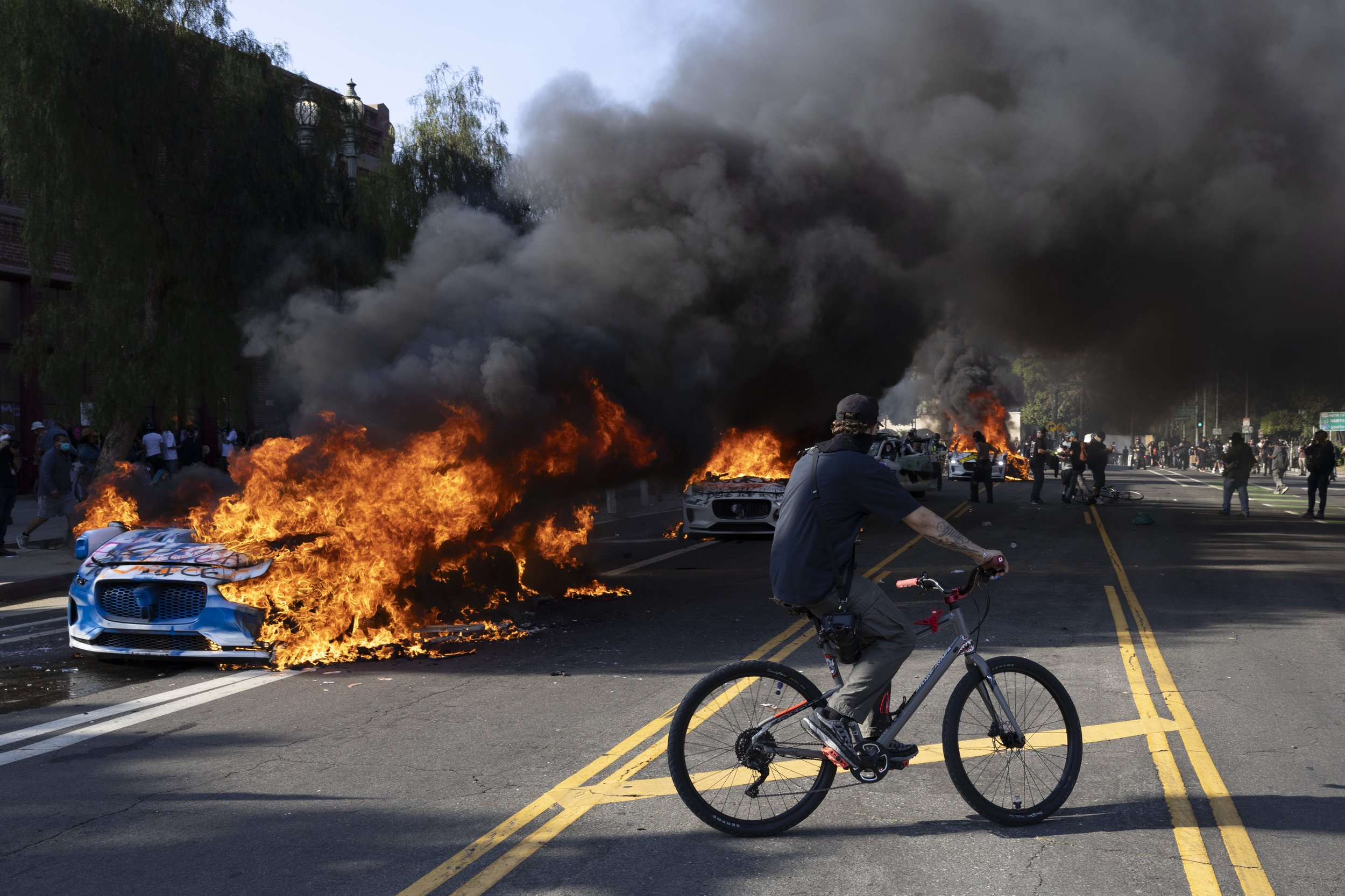 A street scene during a protest or riot with burning vehicles and heavy black smoke, with a person riding a bicycle in the foreground.