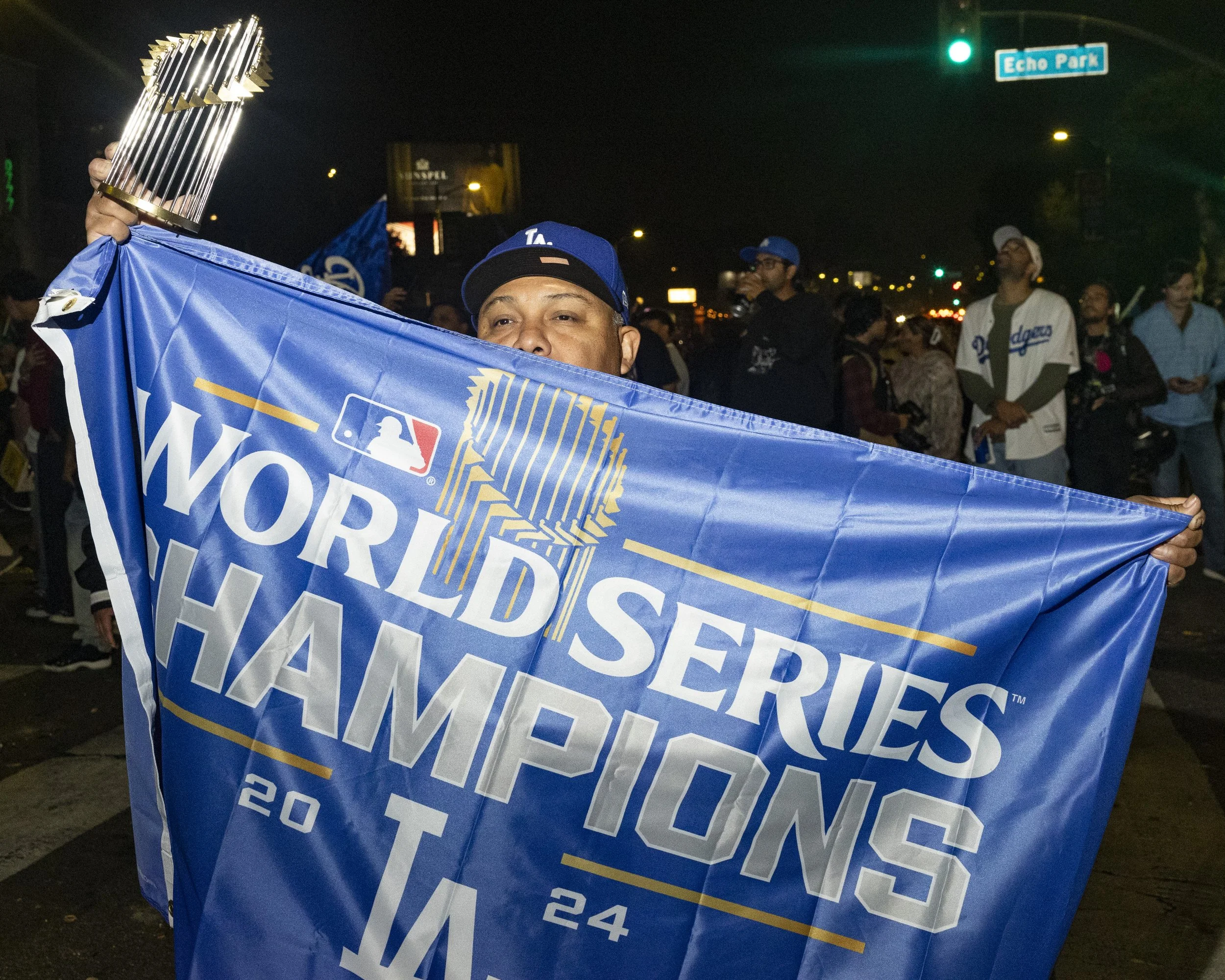 A person holding a Los Angeles Dodgers World Series Champions 2020 flag at night during a celebration parade, with a crowd and street sign in the background.
