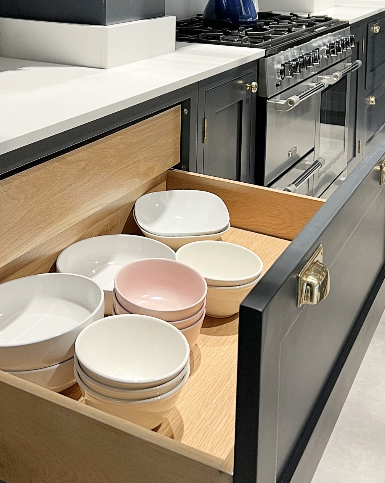 Open kitchen drawer with neatly stacked white and pink ceramic bowls, beside a stove with a kettle on top.