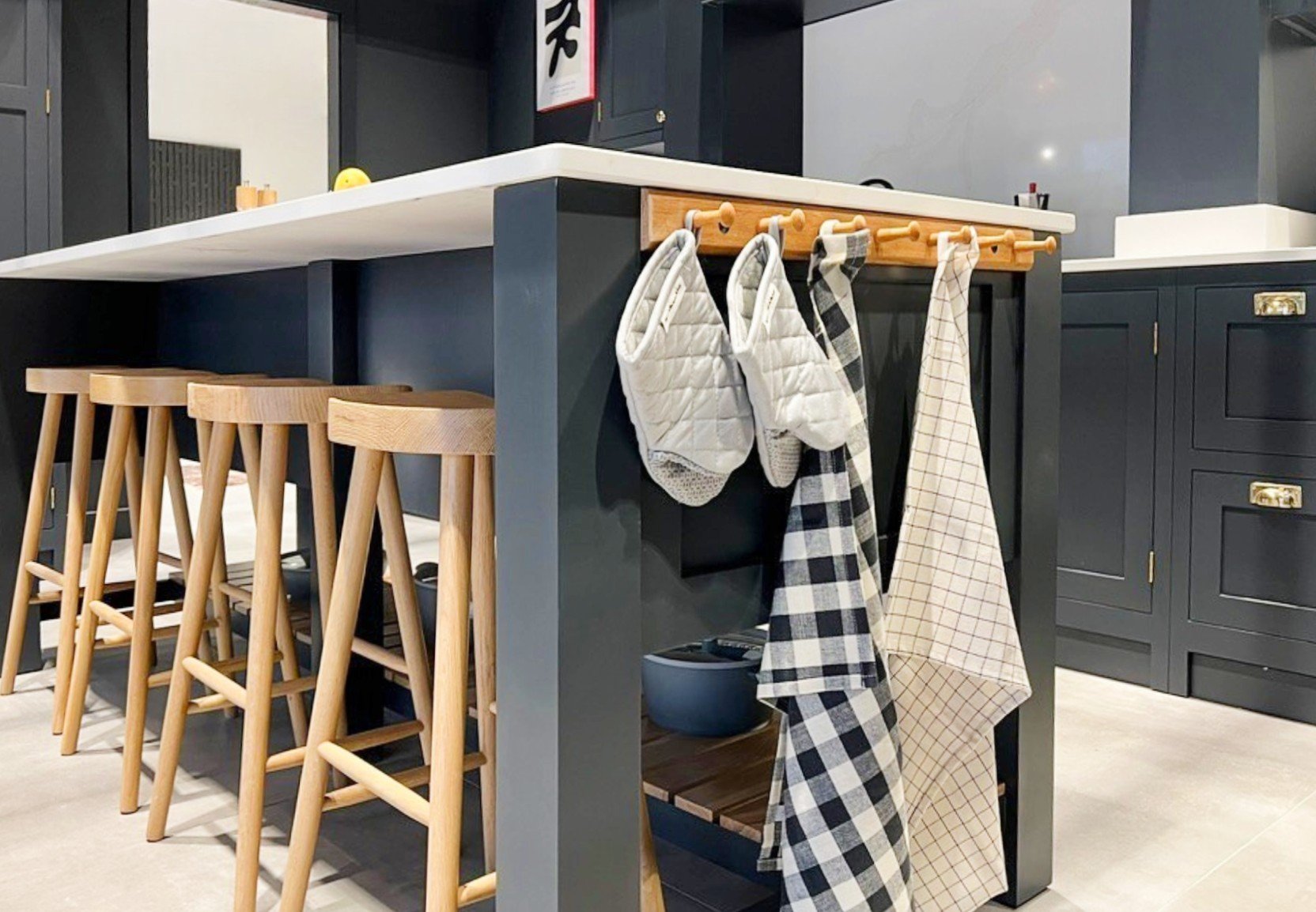 Modern kitchen with a white countertop, wooden bar stools, and dark cabinetry. Oven mitts and checkered kitchen towels hang on a wooden bar.