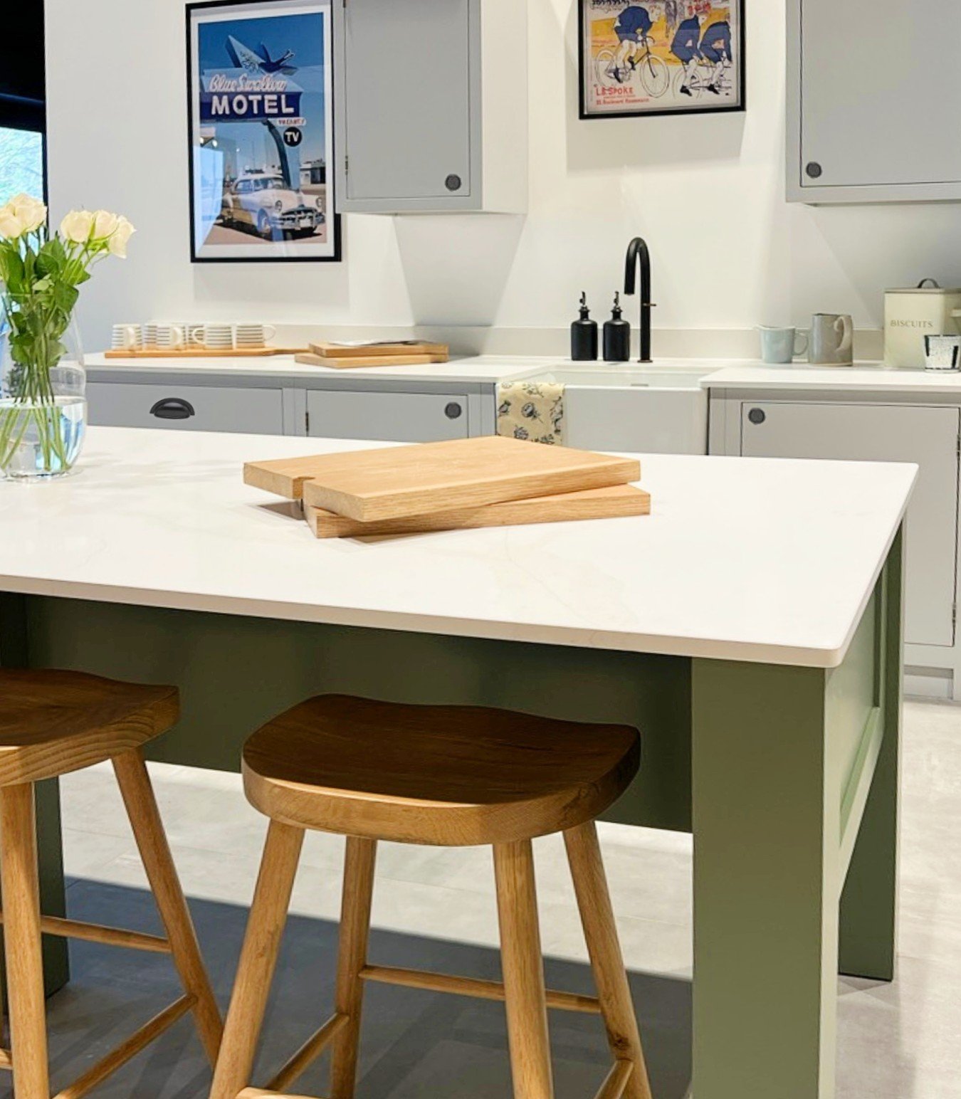 Modern kitchen with wooden stools and cutting board, white countertop, green island, wall cabinets, retro posters, flowers in a vase, and mugs.