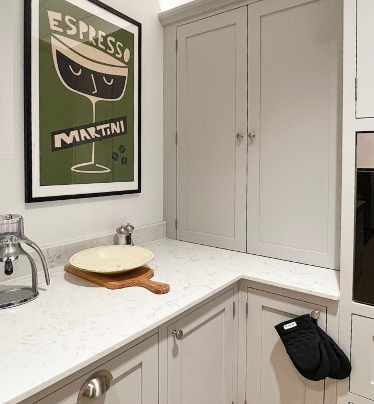 Small corner kitchen area with white cabinets, marble countertops, and an espresso machine. A framed "Espresso Martini" poster hangs on the wall. A wooden cutting board and plate are on the counter. Oven mitts are hanging from a cabinet.