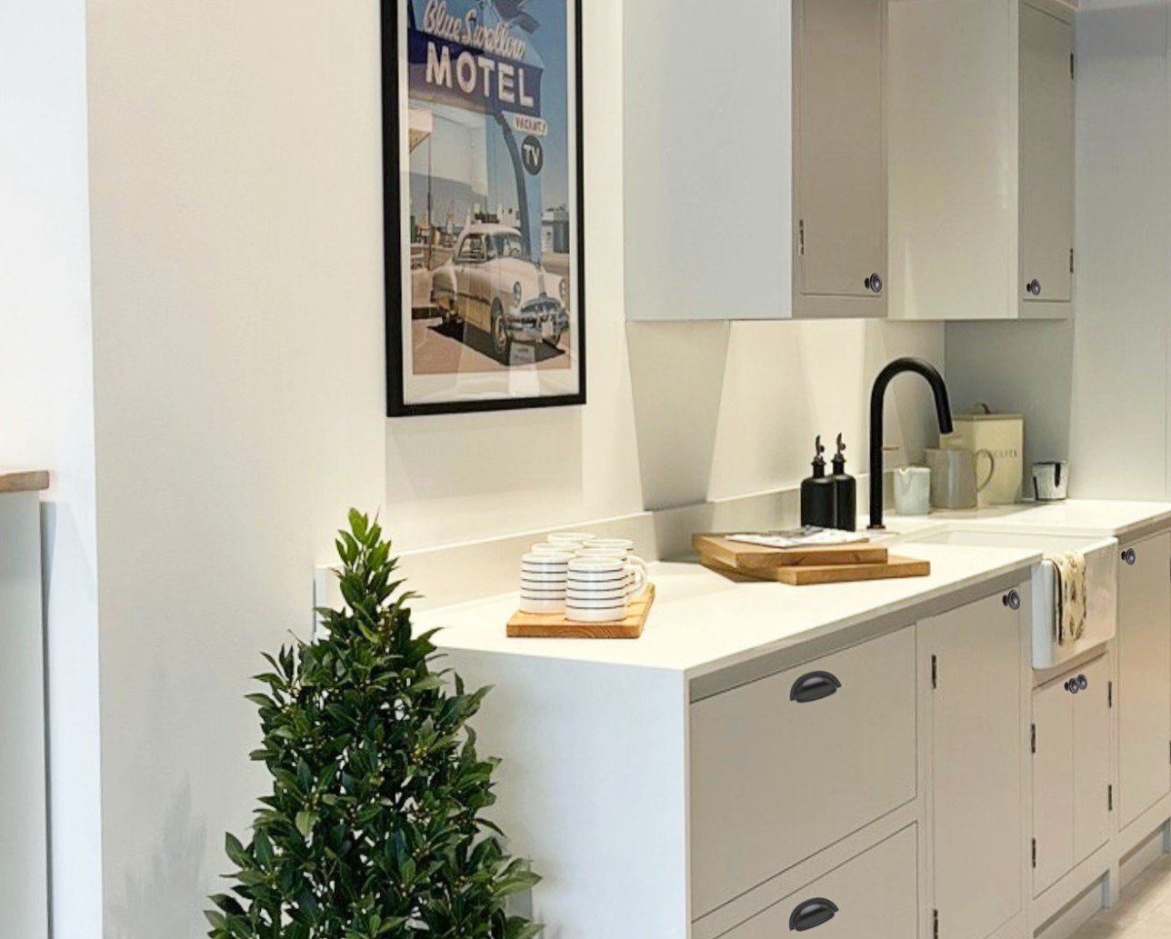 Modern kitchen with white cabinets and a black faucet, decorative plant, vintage motel poster, stacked white cups on a wooden tray, and countertop accessories.