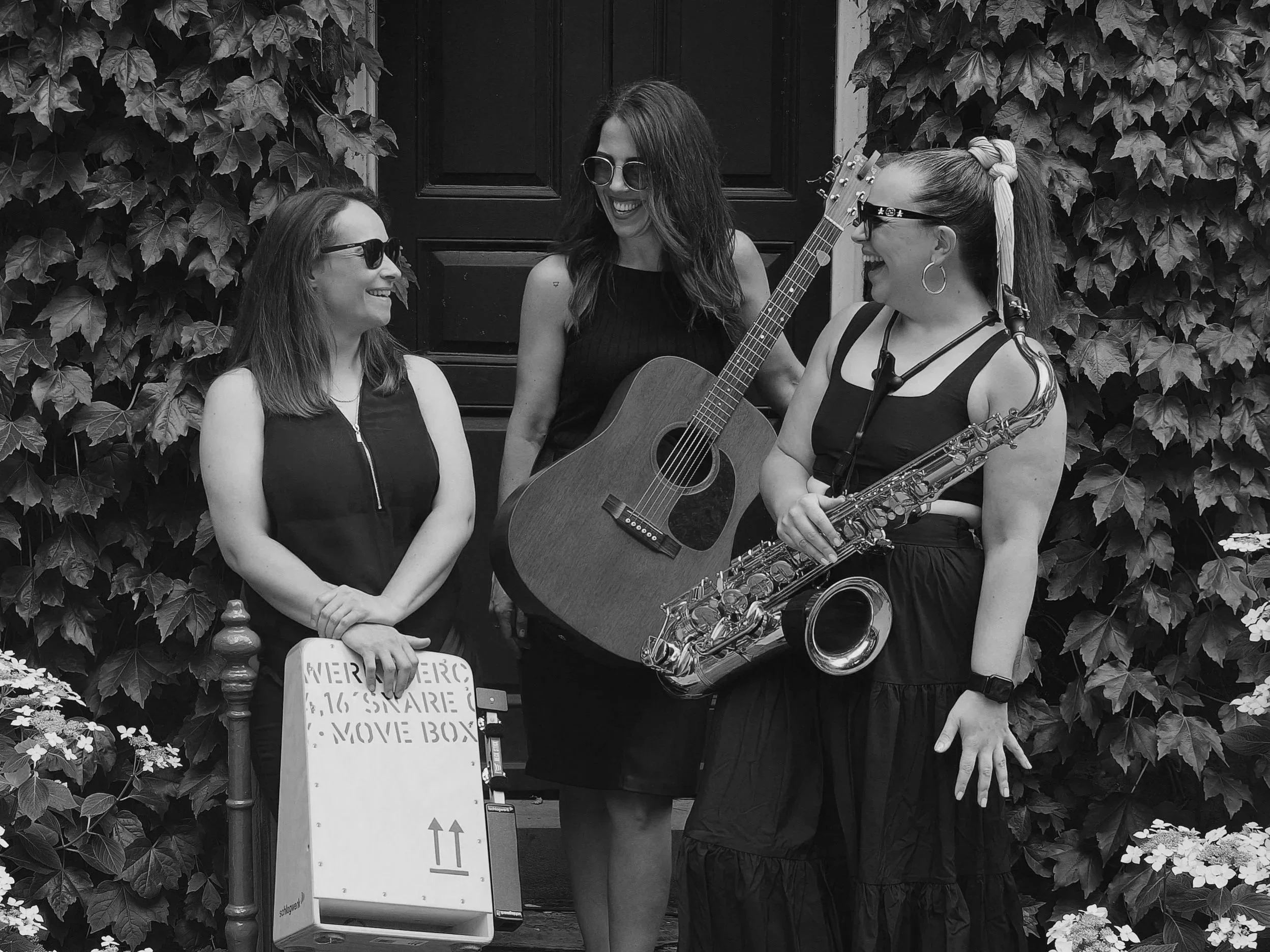 Three women in casual clothing and sunglasses are standing in front of a dark door with leafy vines, laughing and talking. One woman is holding a guitar and another is holding a saxophone.