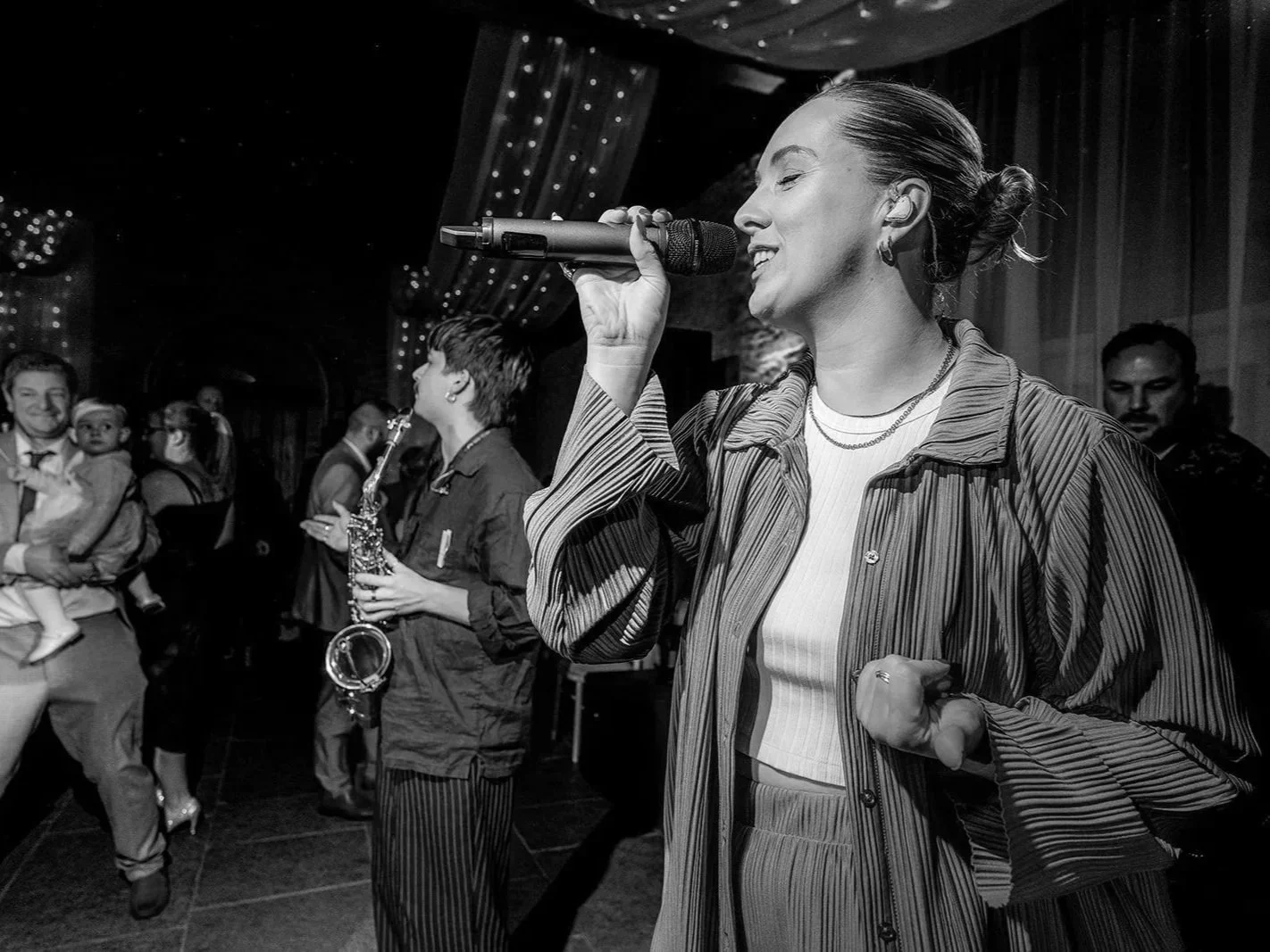 A woman singing into a microphone at a party with people dancing and socializing in the background.