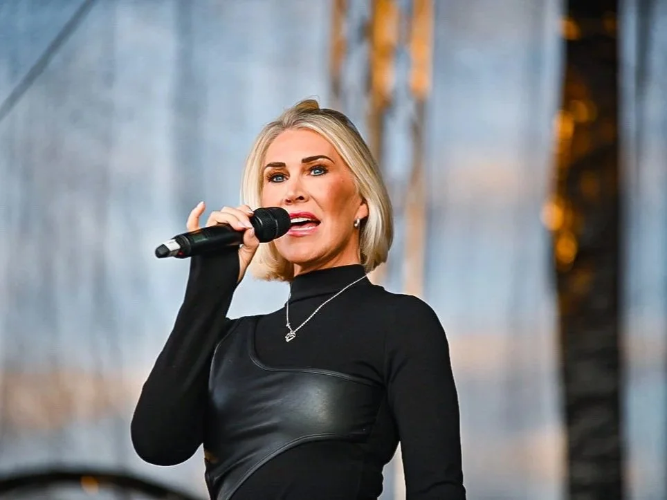 A woman with blonde hair and blue eyes singing into a microphone on stage.