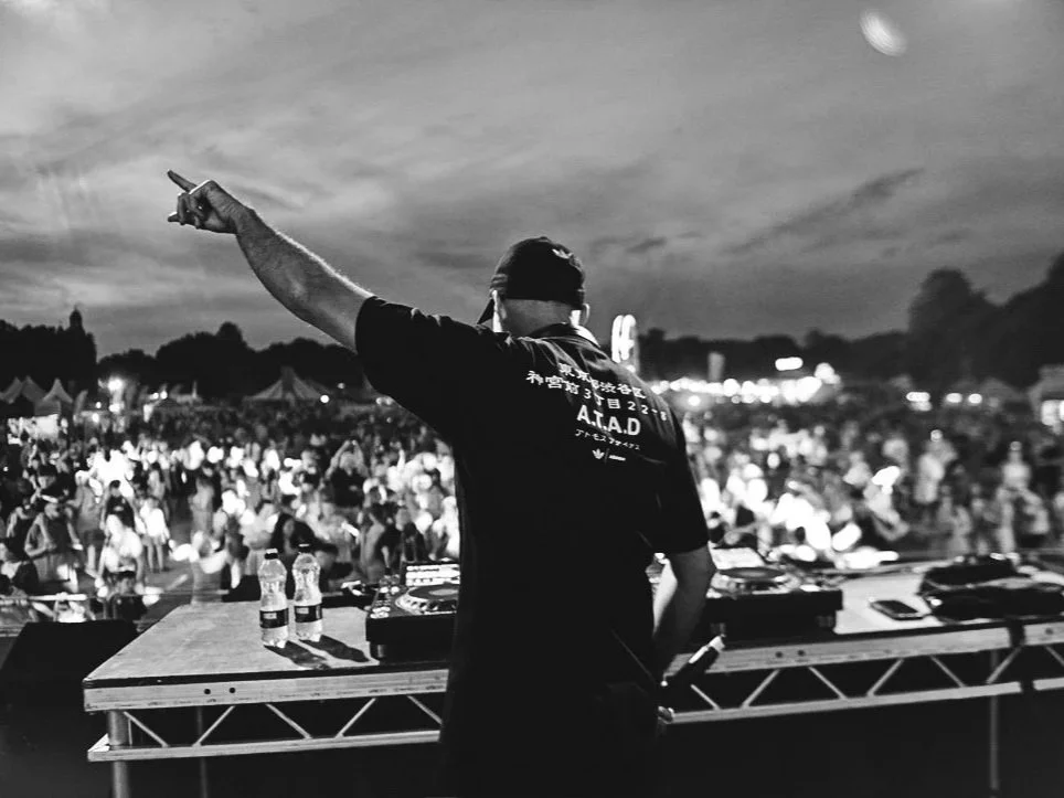 A DJ stands at a table with turntables, raising one hand with a peace sign at an outdoor music festival during dusk, with a large crowd in the background.