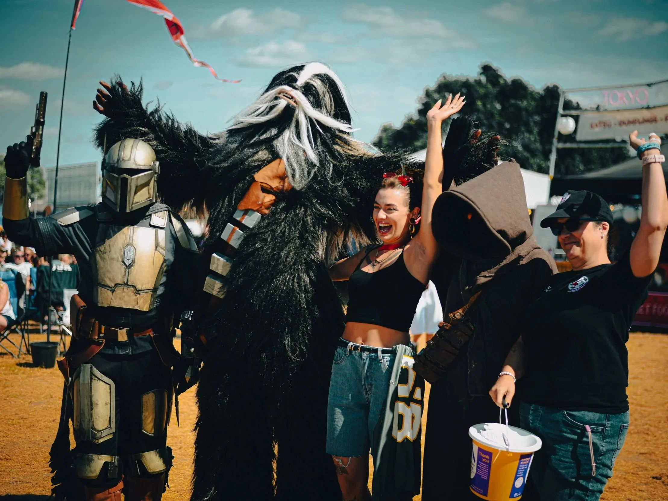 People celebrating at an outdoor event, with one person dressed as Chewbacca from Star Wars and another person wearing a Stormtrooper costume, amidst smiling and cheering crowd.
