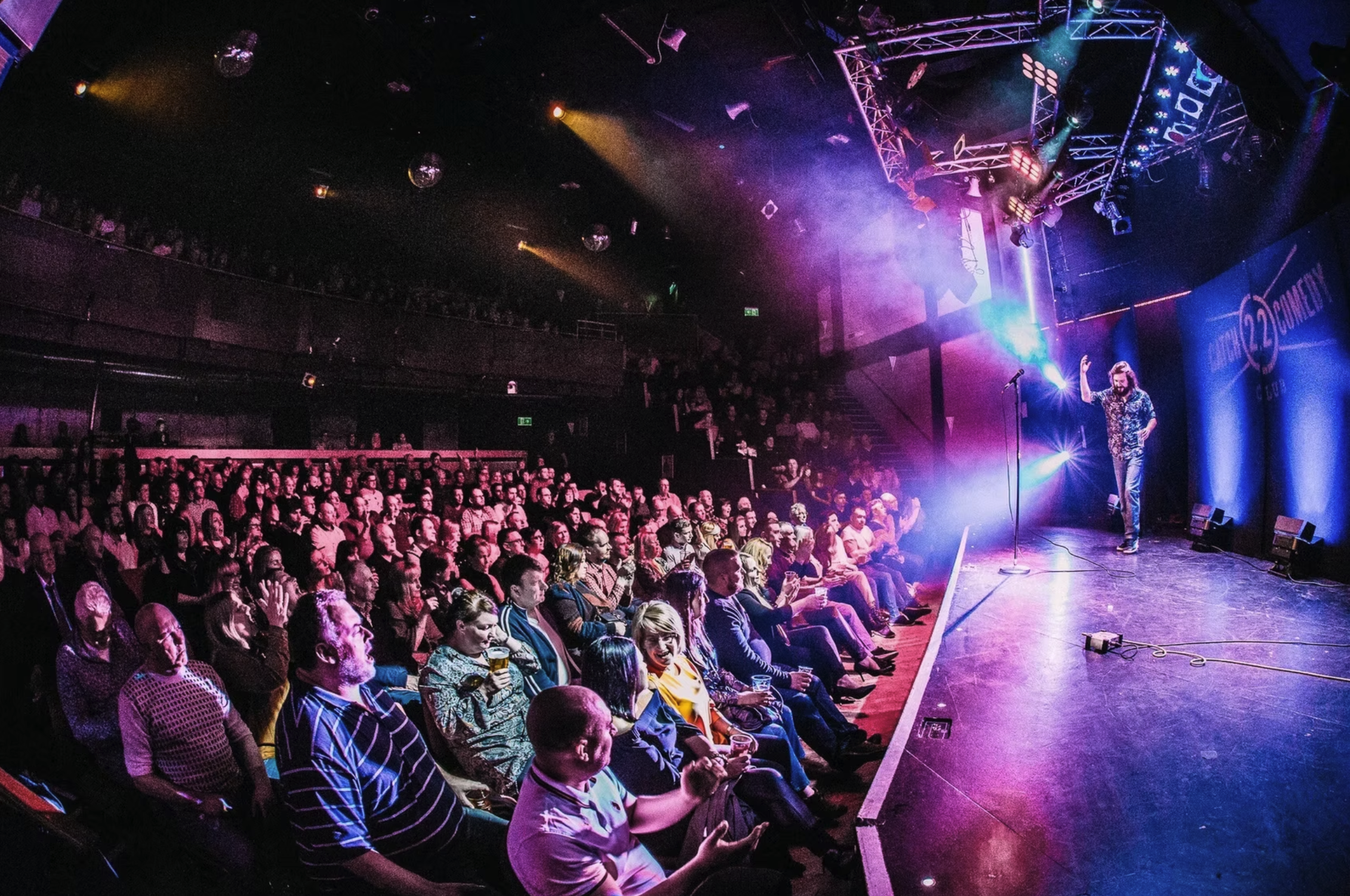Audience watching a comedy performance on stage in a nightclub with colorful lighting and a performer with long hair wearing a patterned shirt. The stage has a microphone and a backdrop with the text '22 Comedy'.