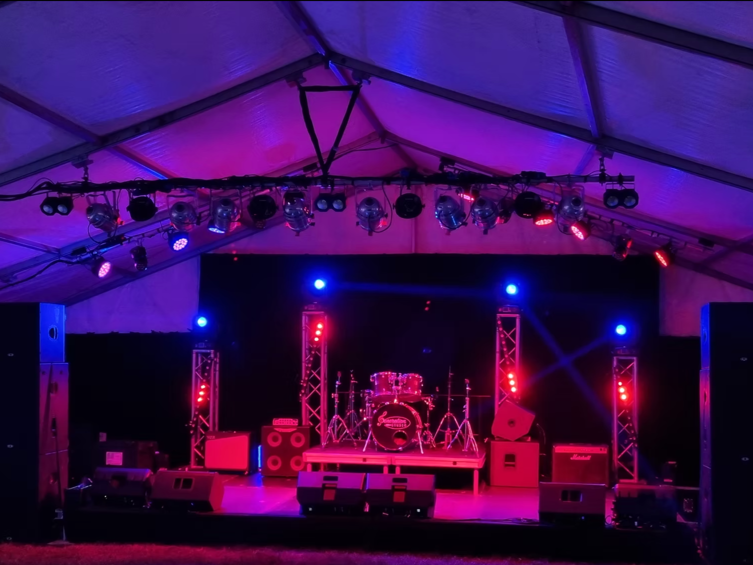 Empty stage with drum set, guitar amplifiers, microphones, and colorful stage lighting in a large tent.