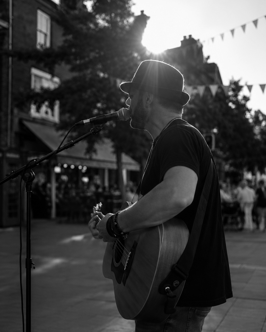 A man wearing a hat playing an acoustic guitar and singing into a microphone outdoors in front of a crowd with bunting decorations, backlit by the setting sun.