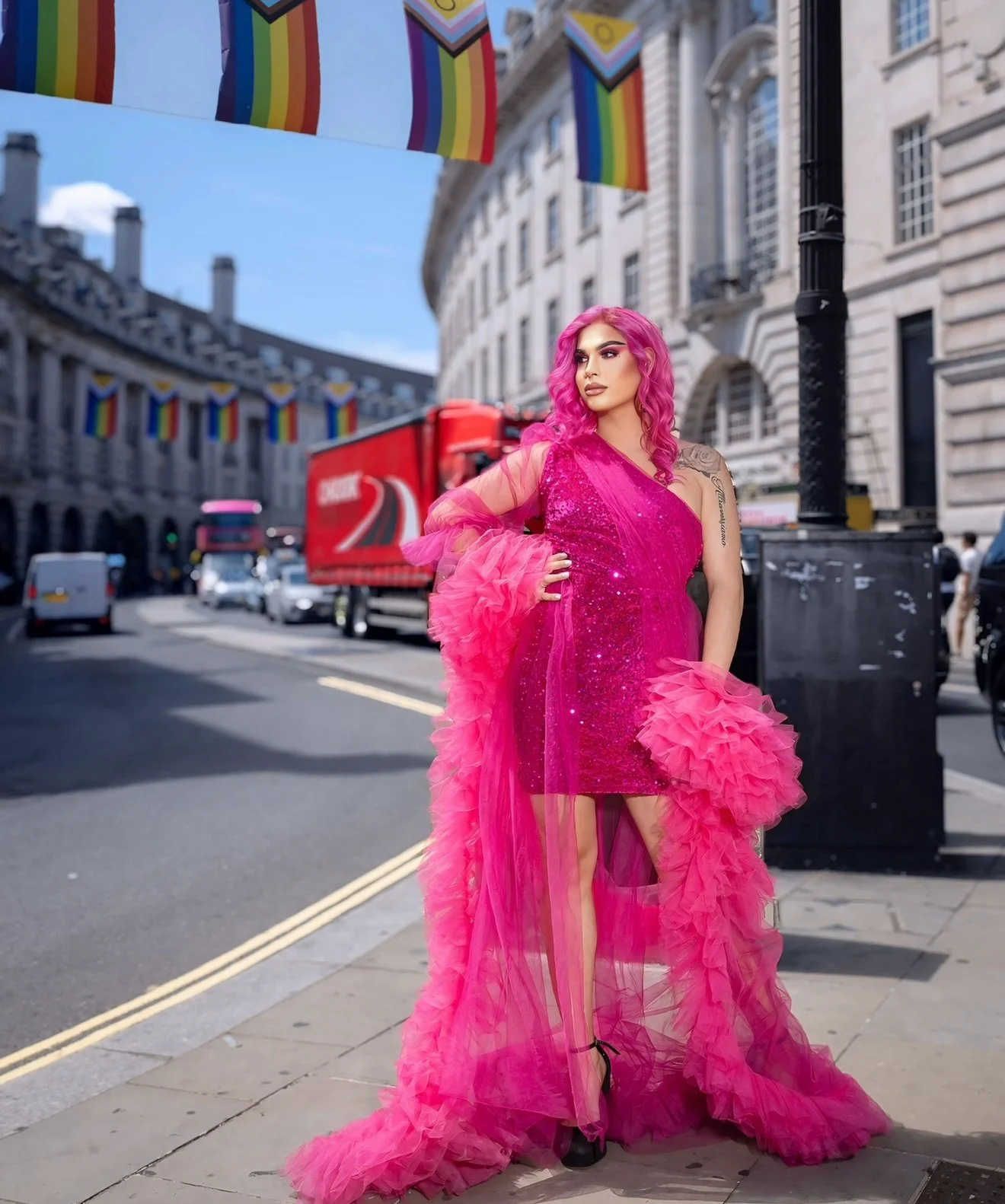 A drag artist with pink hair wearing a vibrant pink dress with ruffled tulle details, standing on a city sidewalk with colourful flags hanging above on a sunny day.
