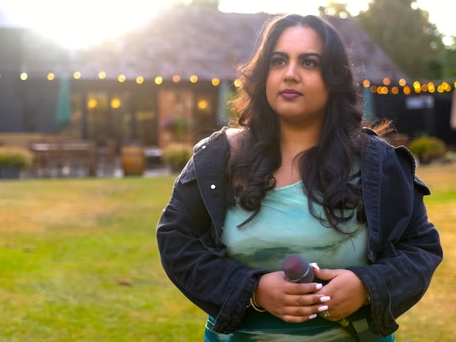 A woman with dark hair holding a microphone outdoors at sunset, with a house and string lights in the background.