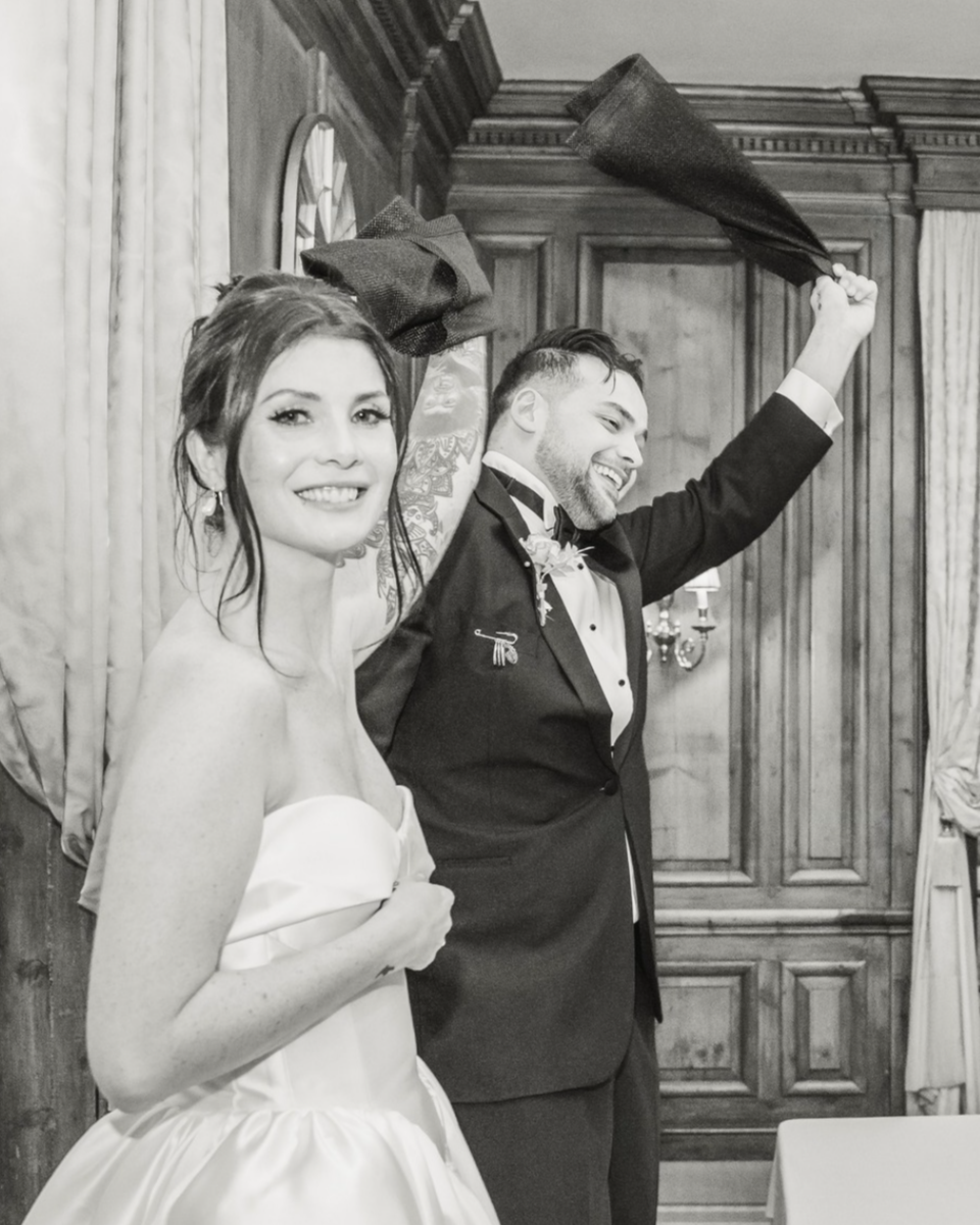 A black and white photo of a bride and groom at their wedding reception, with the groom holding a hat in the air and smiling, and the bride smiling and holding her gown.