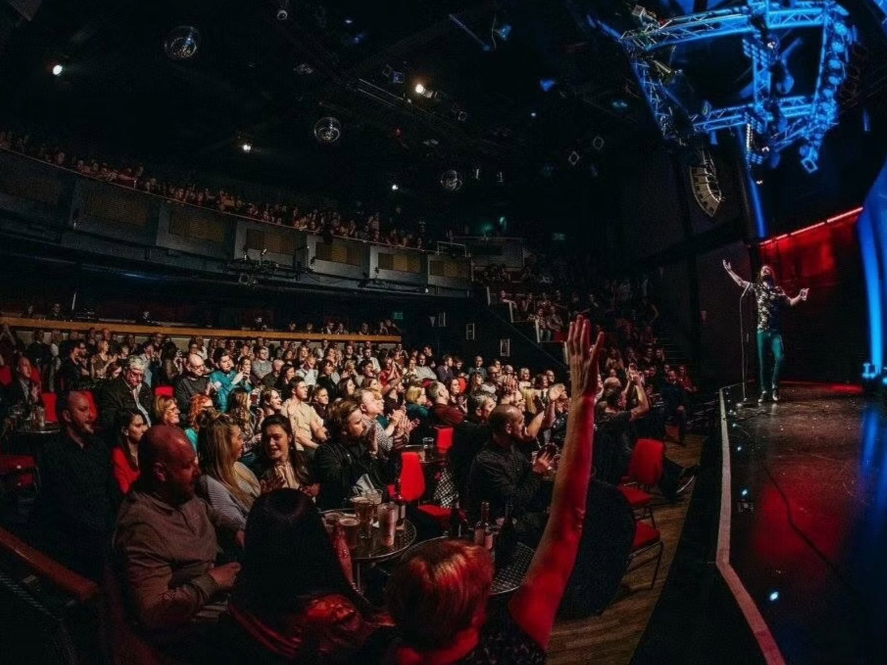 A comedian performs on stage at a comedy club with a large audience clapping and laughing, seated at tables with drinks, in a dimly lit venue with stage lighting and a balcony level.