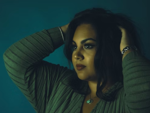 A woman with dark hair and makeup, wearing a green striped top, posing with her hands in her hair against a dark teal background.