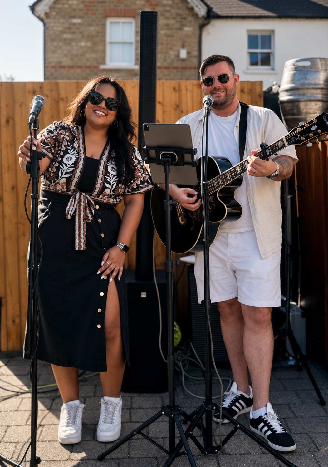 Two musicians perform outdoors, a woman with dark hair, sunglasses, and a patterned top, singing into a microphone, and a man with short hair, sunglasses, playing an acoustic guitar, standing in front of a wooden fence.