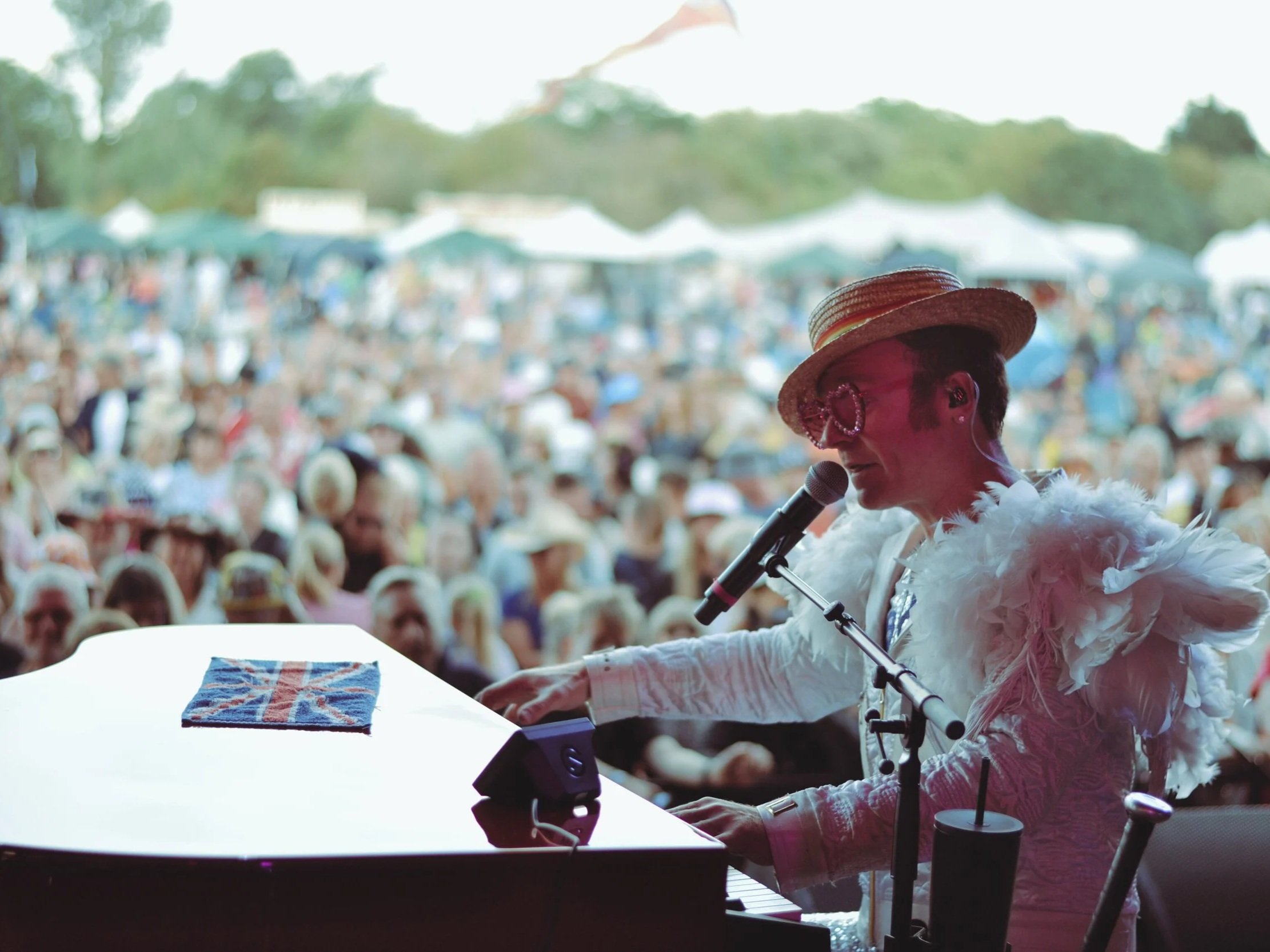 A woman wearing a straw hat and feathered white jacket singing into a microphone at an outdoor event with a blurred crowd and tents in the background.