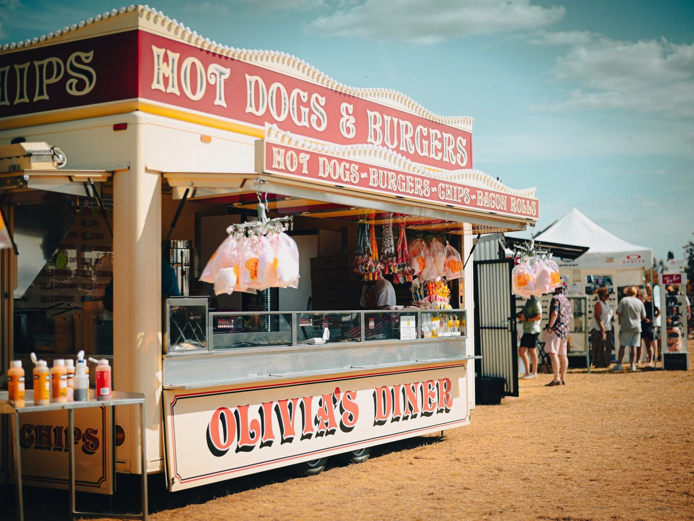 A food stand at an outdoor event selling hot dogs, burgers, chips, and bacon rolls, with a sign displaying the menu. There are people walking in the background and some hanging merchandise.