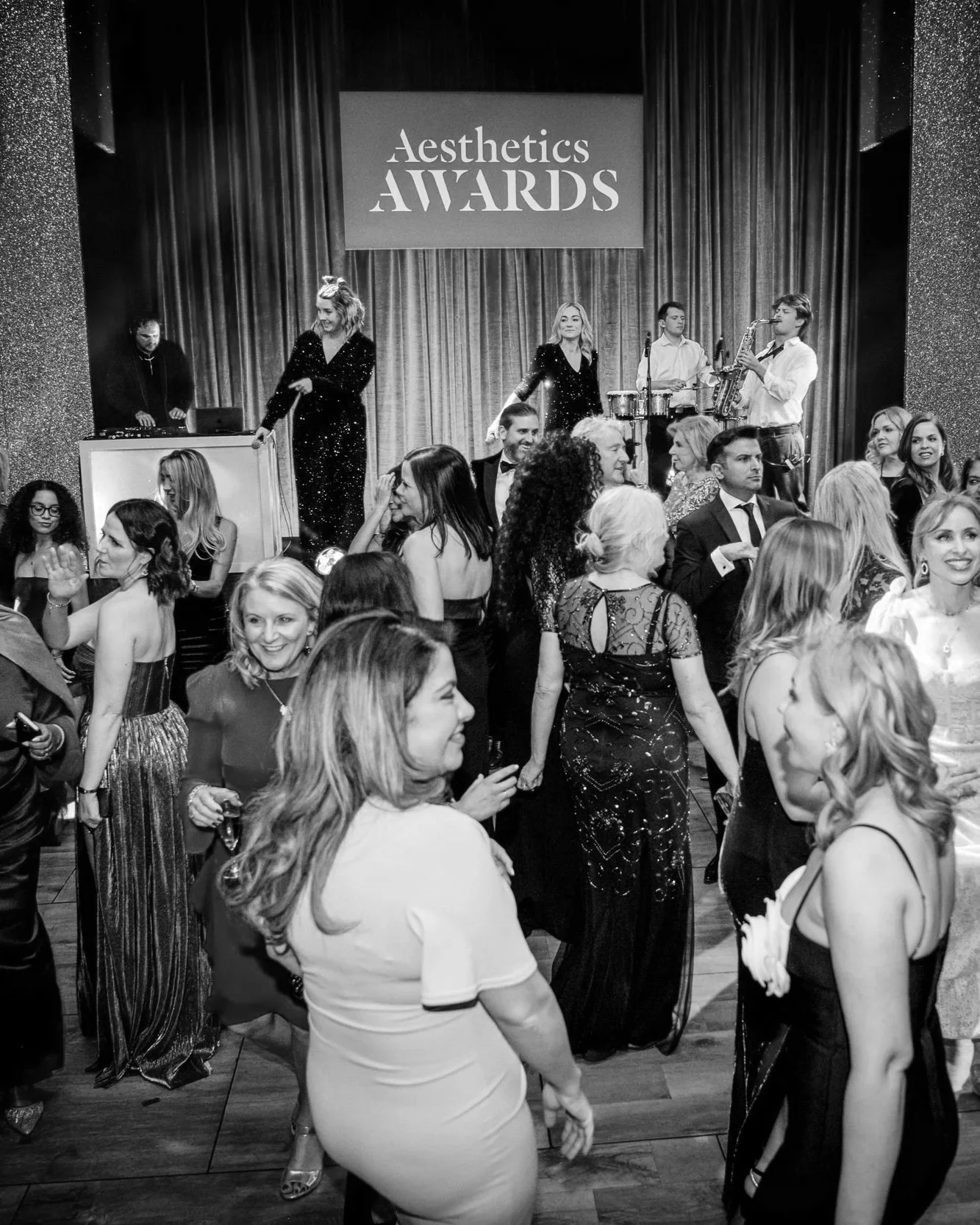 Black and white photo of a lively crowd dancing at an event called 'Aesthetics Awards.' There is a live band playing on stage with a singer and musicians, and a DJ is in the background.