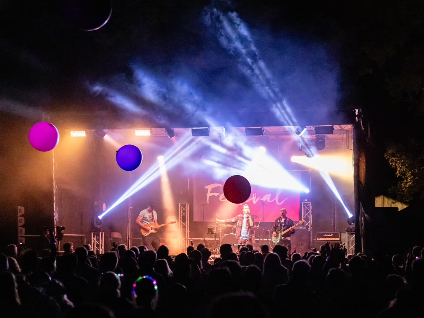 Live music concert with a band performing on stage, audience watching, colorful stage lighting, and large balloons in the crowd