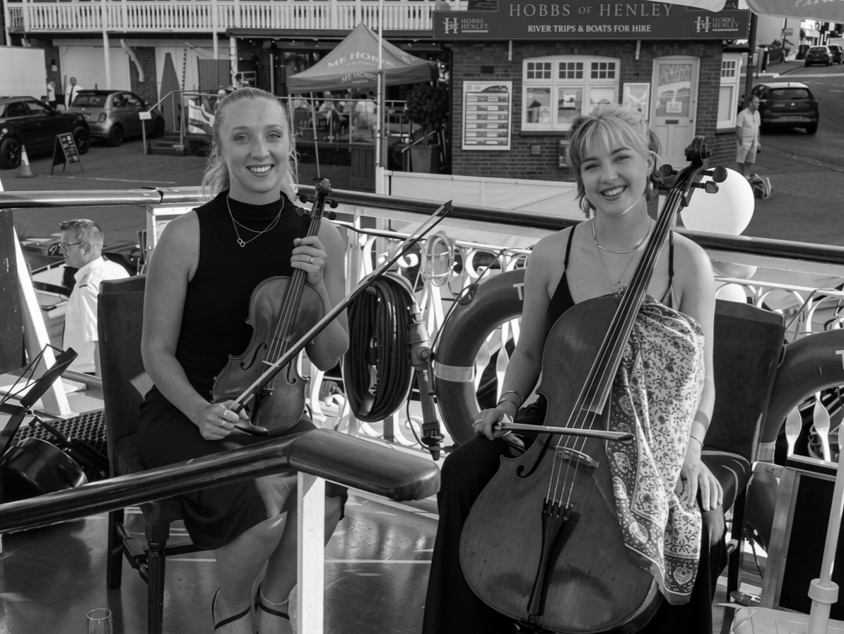 Two young women playing string instruments, one with a violin and the other with a cello, on a boat deck. Sheer smiles on their faces, with a background showing a street, parked cars, and a building with a sign.