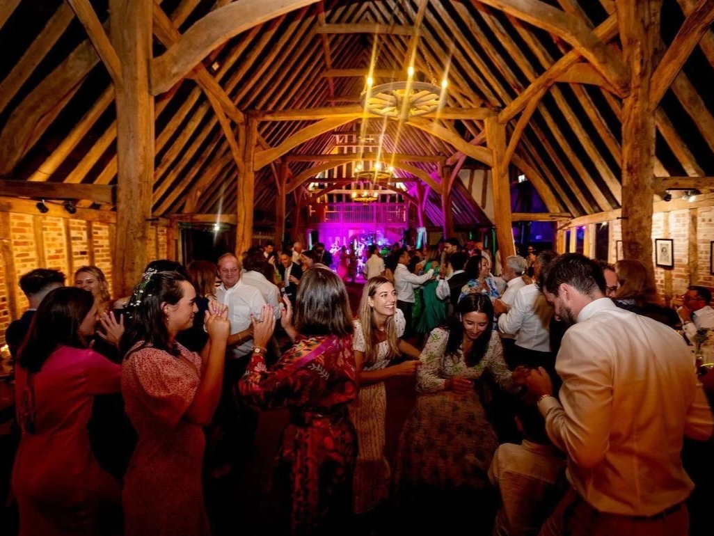 People dancing and socializing inside a rustic wooden event hall with exposed brick walls and warm lighting.