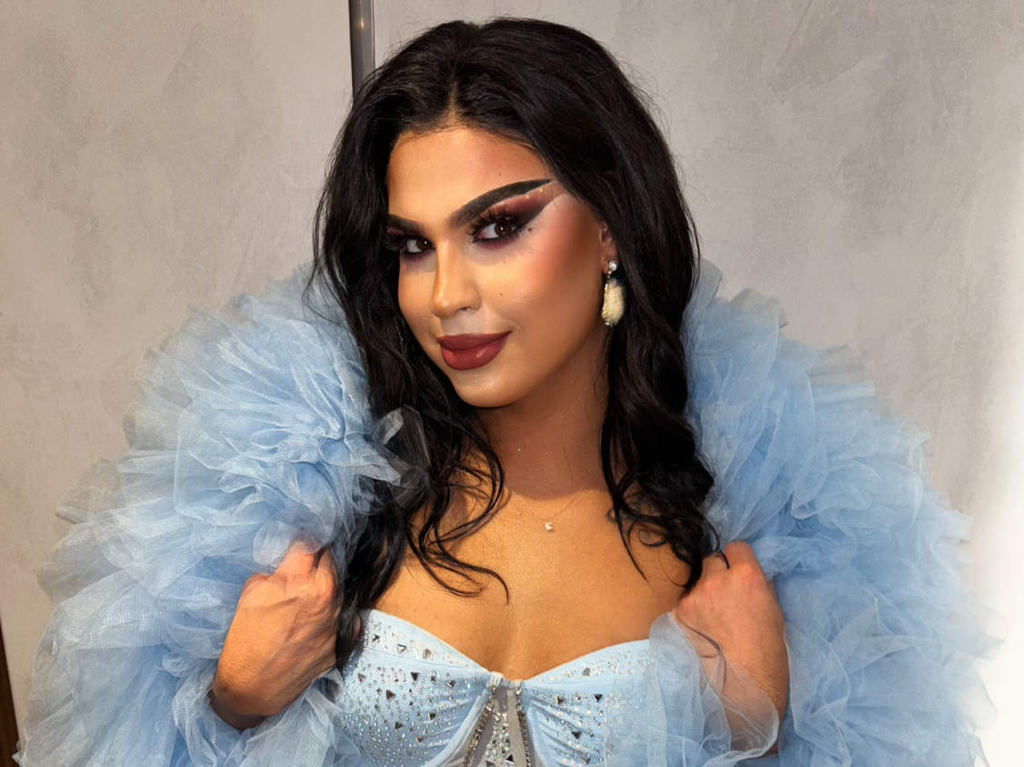 A woman with dark wavy hair dressed in an elegant light blue gown with ruffled sleeves, makeup, and jewelry, posing indoors.