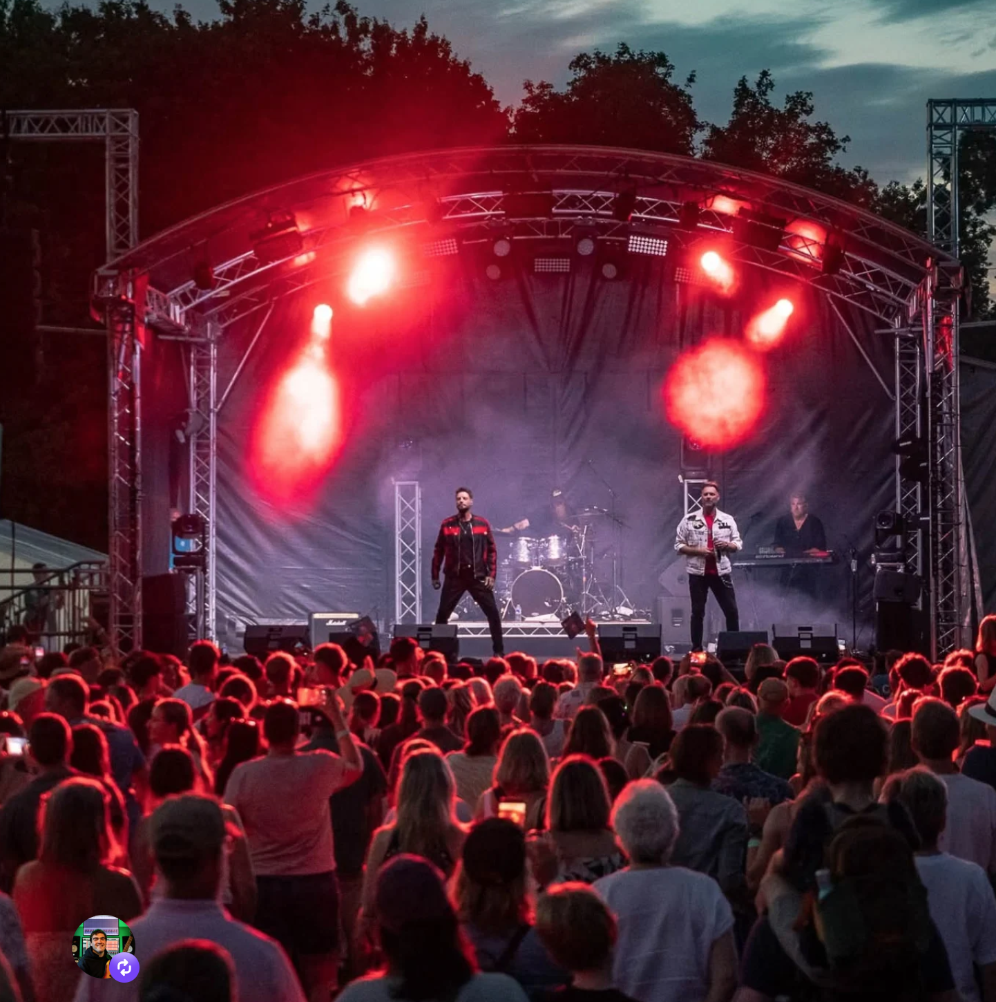 A live outdoor concert with a band performing on stage, illuminated by red lights, in front of a large audience at dusk.