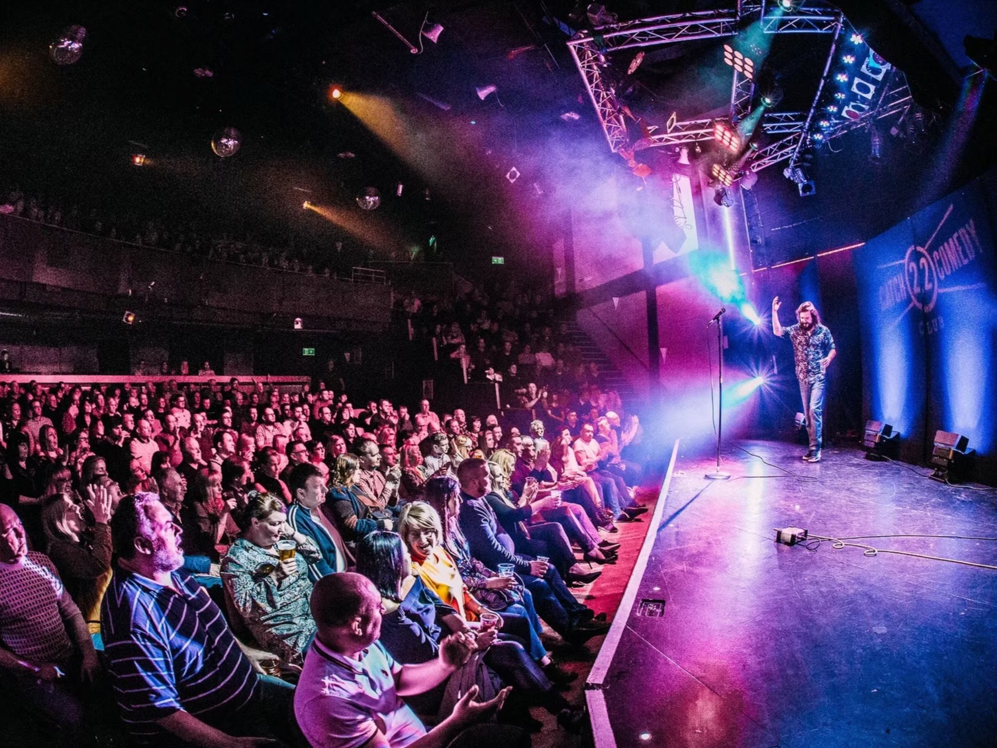 A comedian performs on stage at a comedy club, with a large audience watching and some holding drinks, under colorful stage lighting.