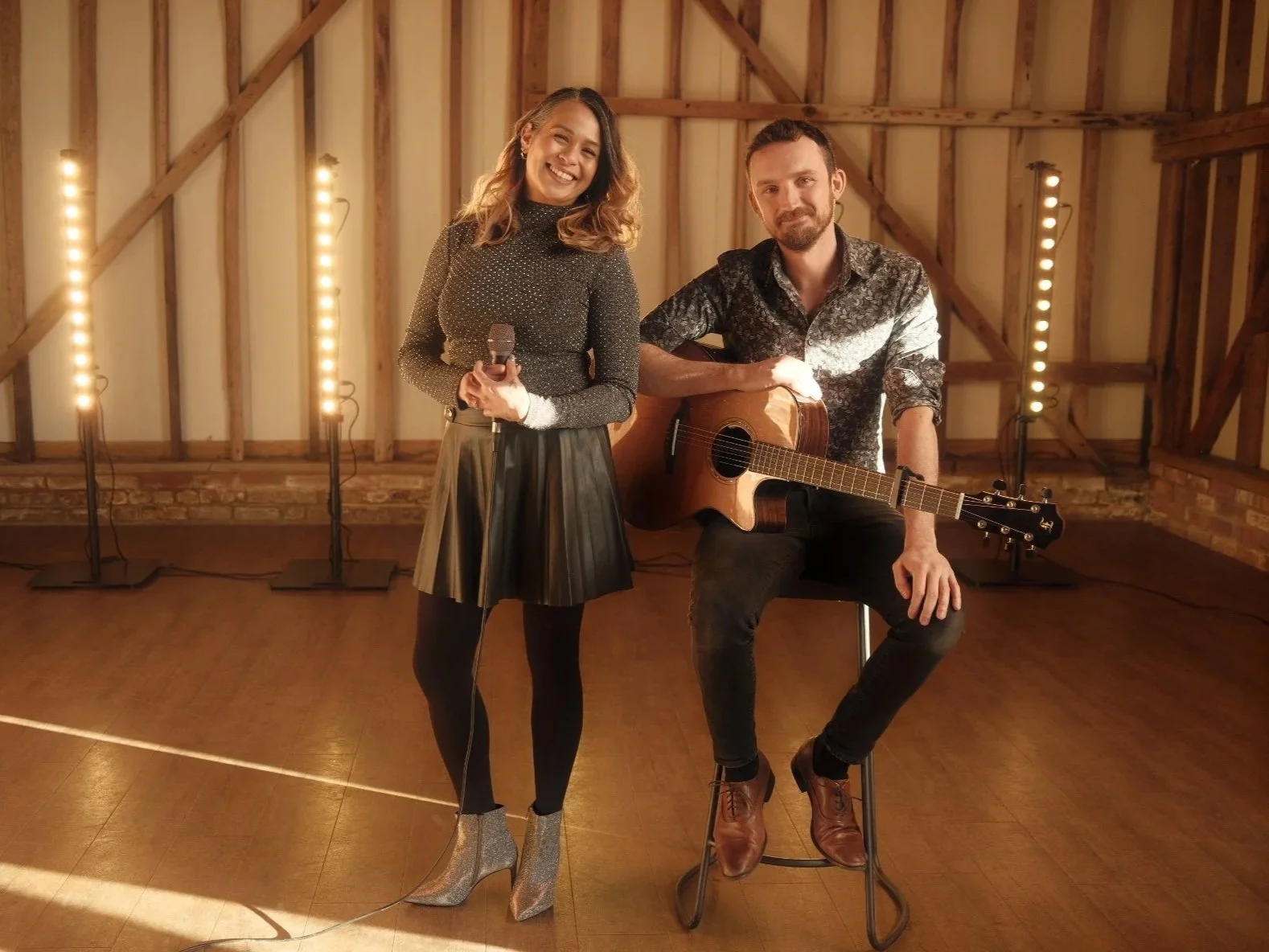 A woman holding a microphone and a man playing an acoustic guitar, seated on a stool, in a warmly lit room with wooden walls and floor.