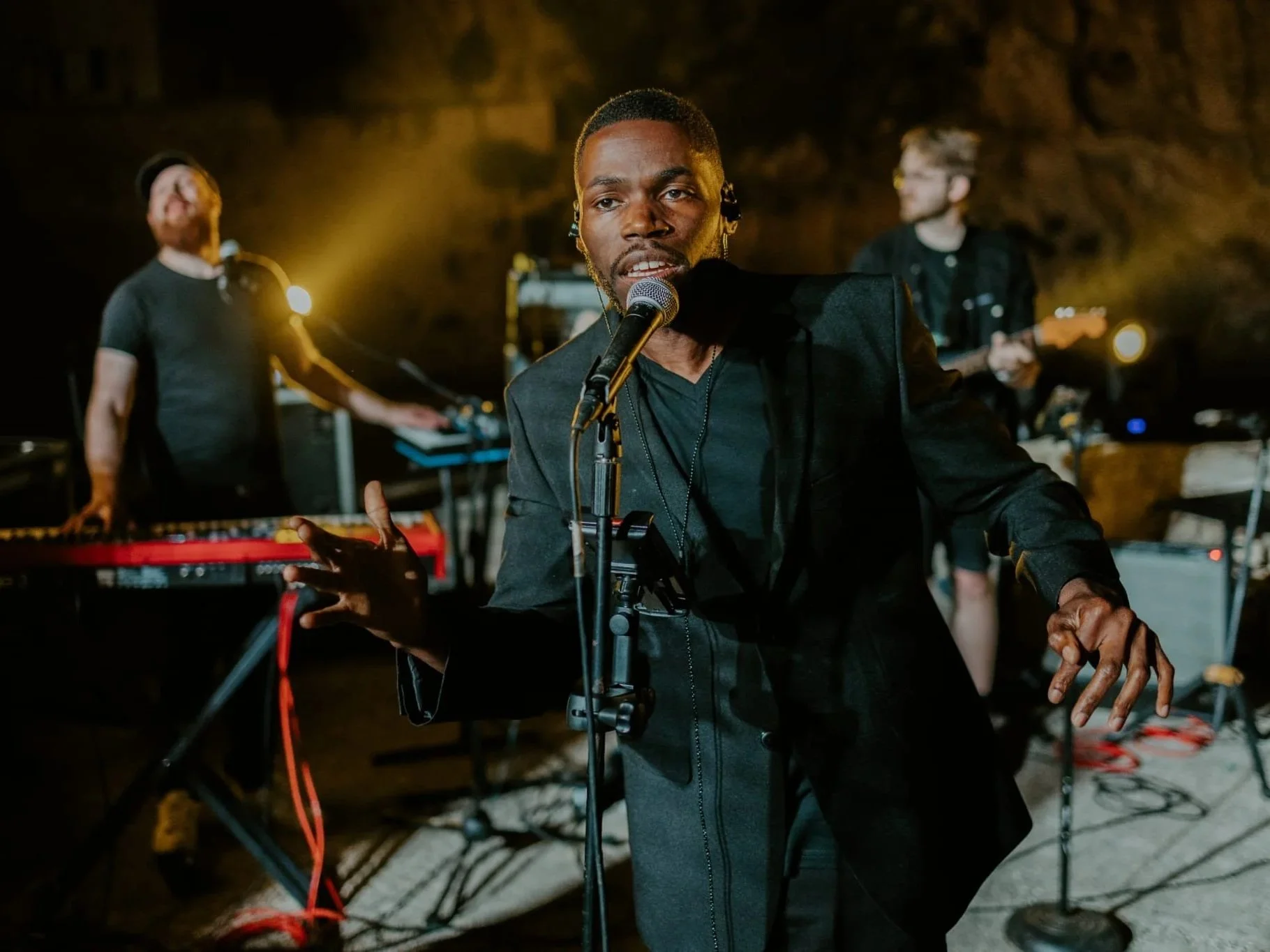 A singer performing live with a band behind, with keyboard and guitar, in a dimly lit venue.
