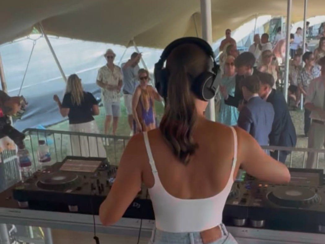 A woman DJ wearing headphones performs at an outdoor event with a crowd dancing and socializing under a tent.