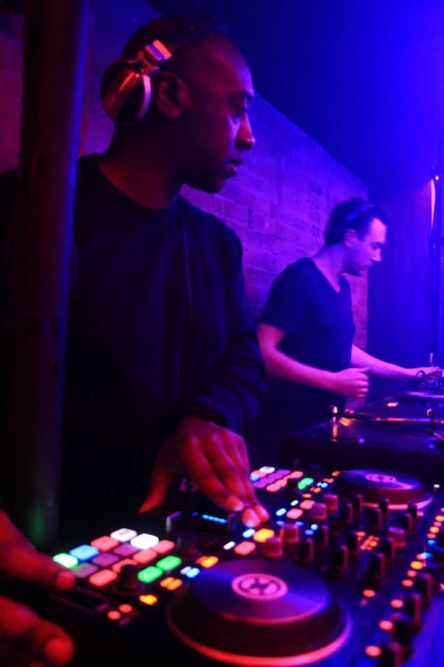 A DJ with headphones focused on mixing music at a club with neon lights, alongside a woman playing a turntable.