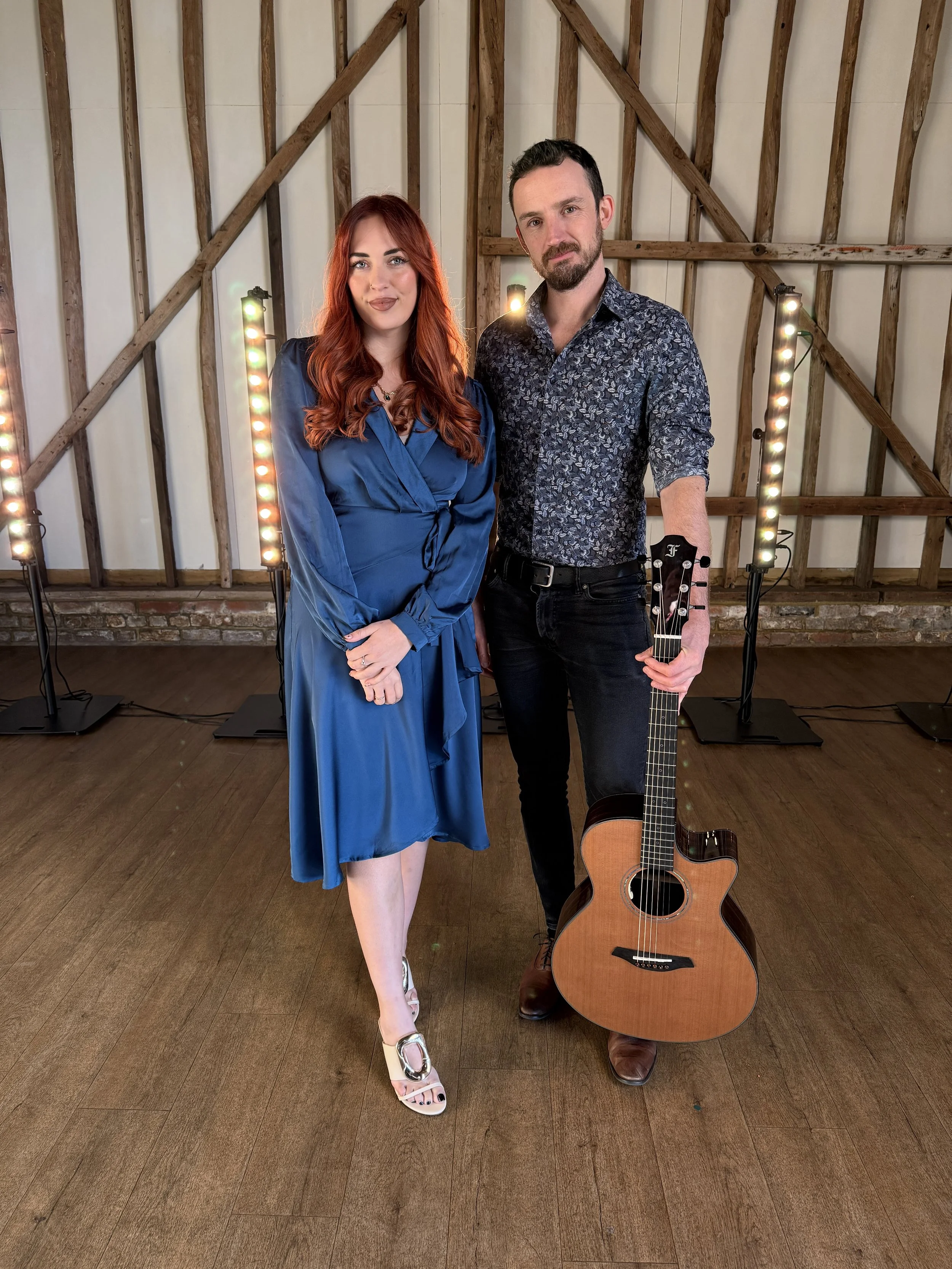 A man and woman standing together indoors in front of wooden wall with stage lights, the woman wearing a blue dress and the man holding an acoustic guitar.