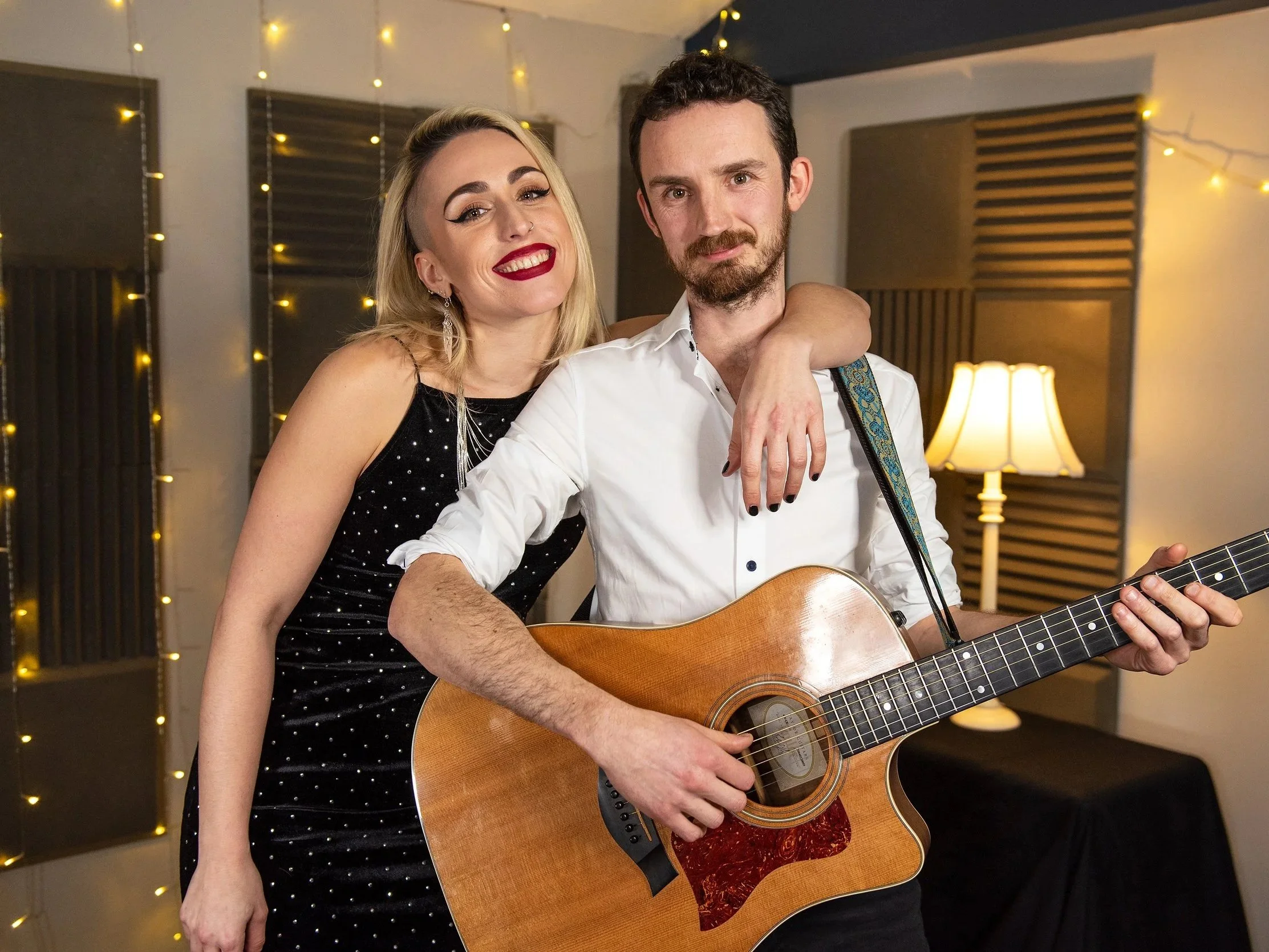 A smiling woman with blonde hair and red lipstick leans on a man with a guitar, in a room decorated with string lights and a table lamp.