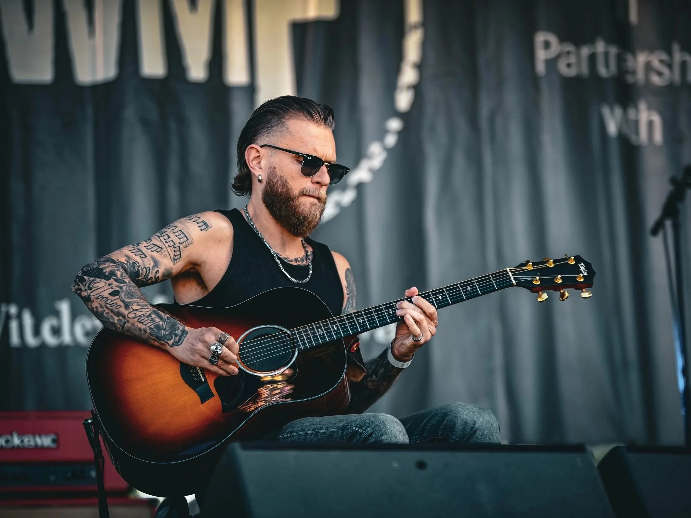 A man with long hair and tattoos playing an acoustic guitar, wearing dark sunglasses, a black sleeveless shirt, and jewelry, performing on stage with a dark backdrop and some musical equipment visible.